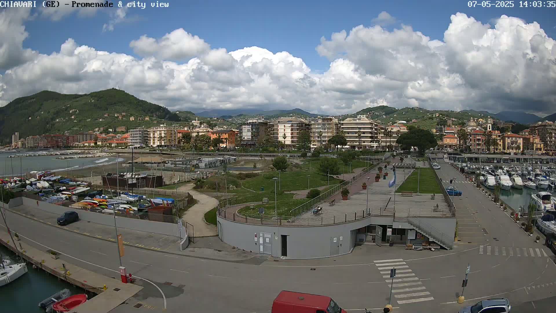 A partly cloudy day reveals a coastal city with a marina filled with boats, a promenade with green spaces, and buildings nestled against a hillside.