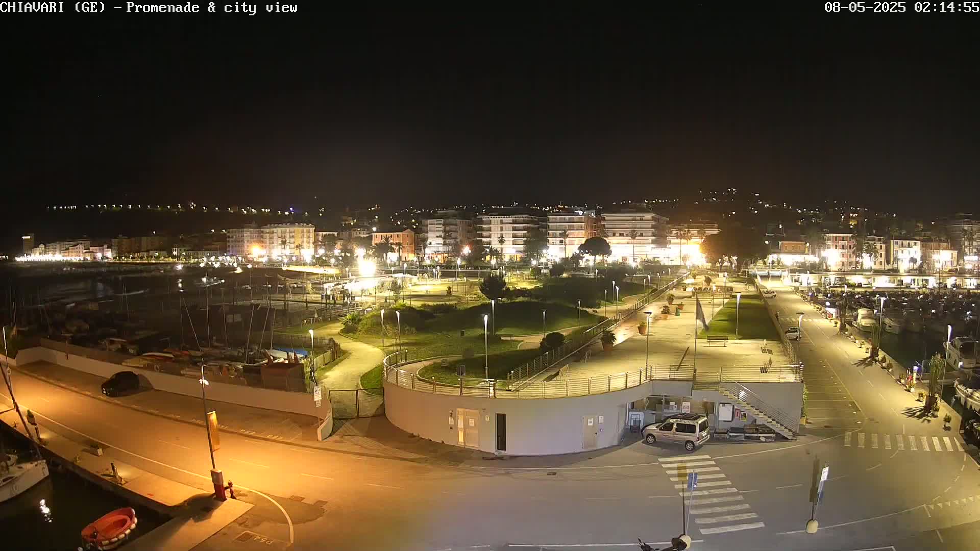 A nighttime, illuminated view of a harbor promenade and city, featuring a marina with boats, a park, and buildings along the waterfront under clear skies.