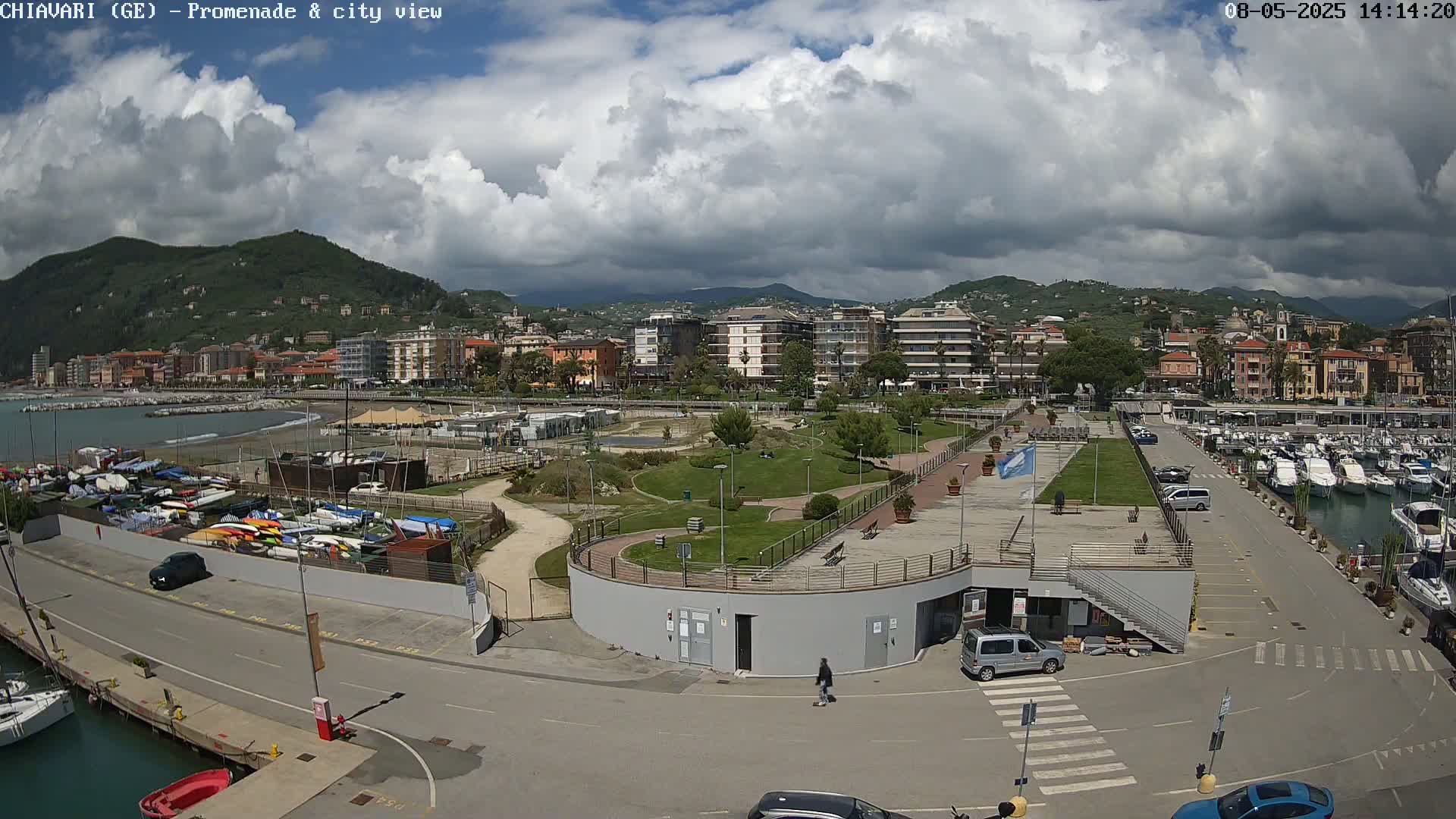 A harbor scene with numerous boats, a promenade, a park, and buildings nestled against a hillside under a partly cloudy sky.