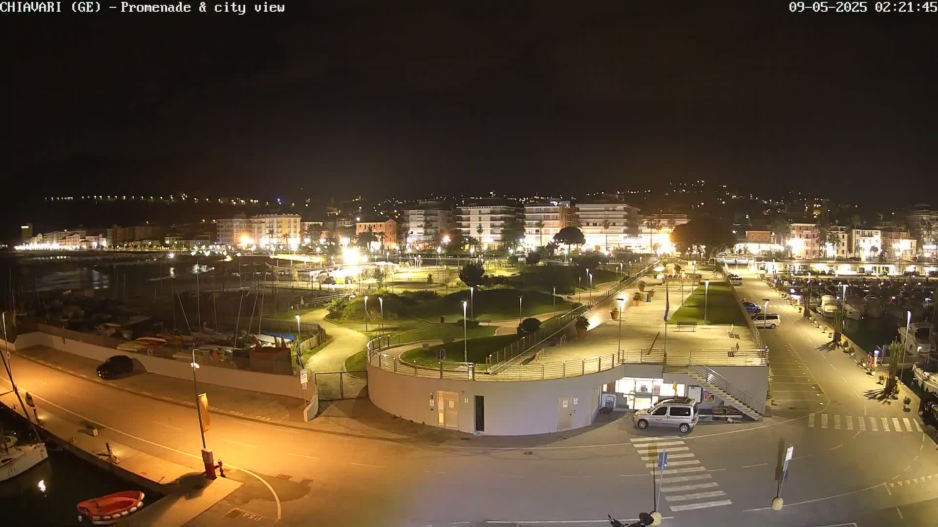A nighttime view of a harbor and city, illuminated with artificial lights, showing a promenade, a marina filled with boats, and buildings along a waterfront.