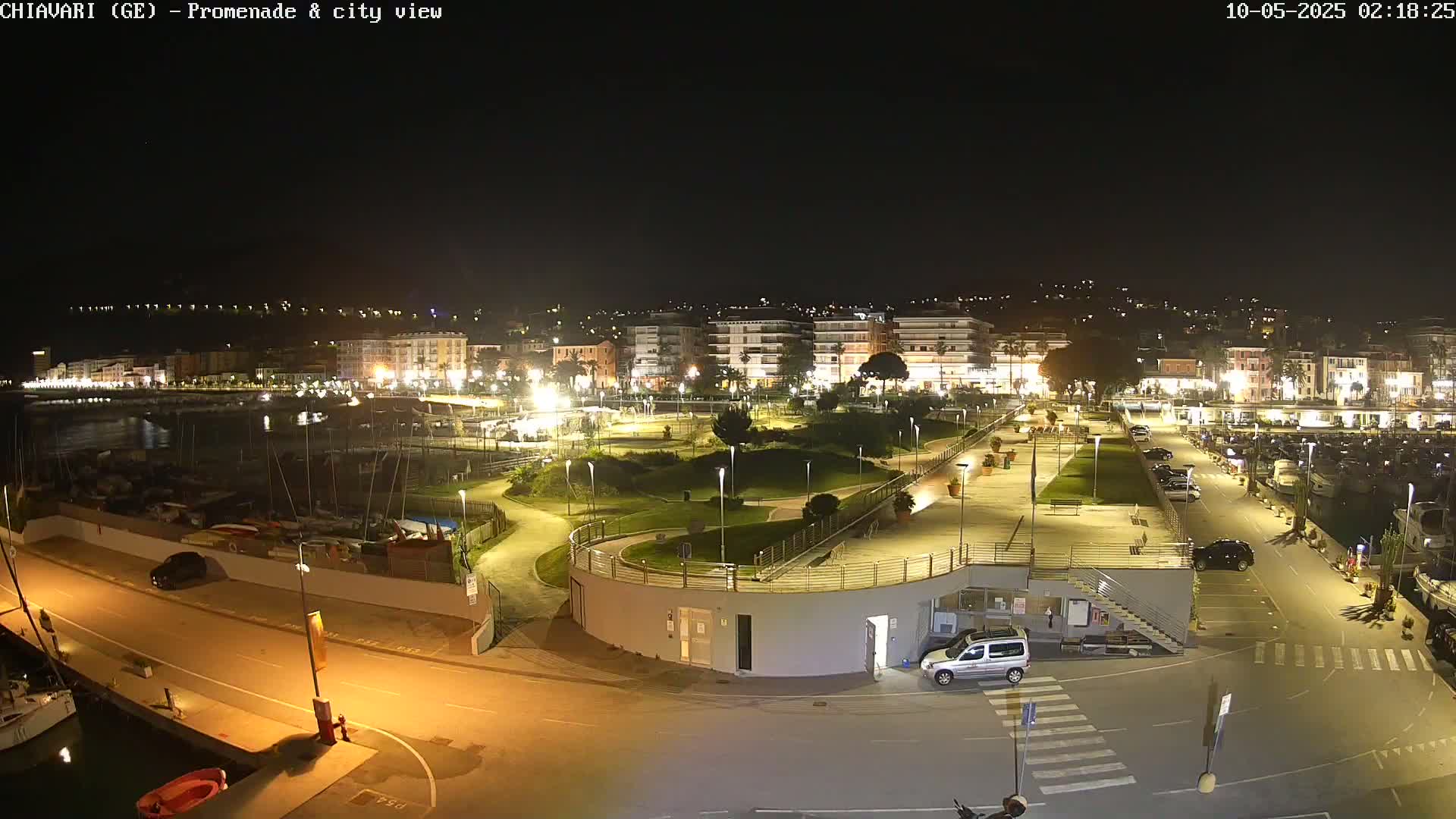 A nighttime view of a waterfront promenade and city, illuminated with streetlights, showing a harbor with boats and a park-like area.