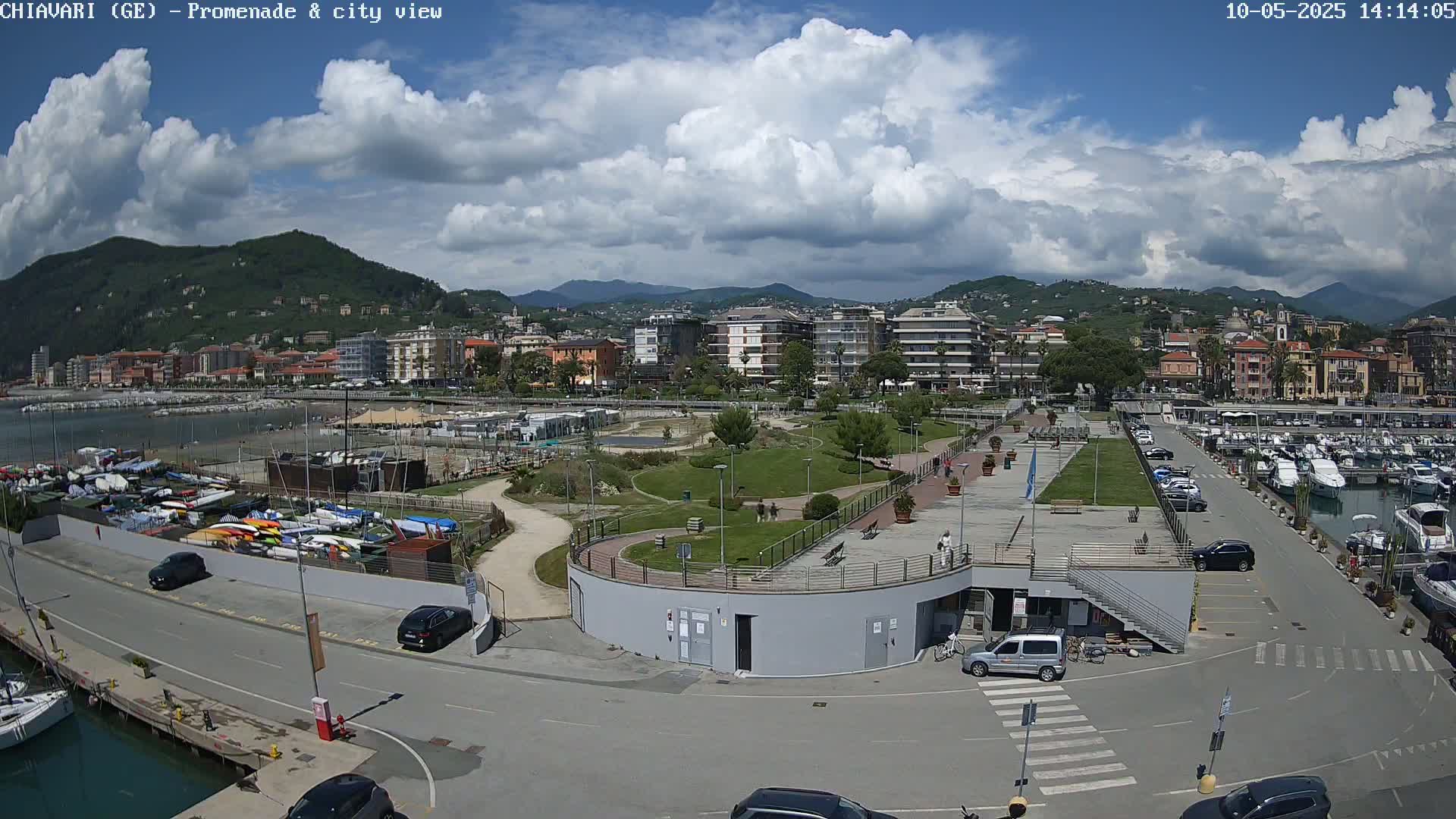 A partly cloudy day overlooks a waterfront promenade with a marina, a park, and a city of buildings nestled against mountains in the background.