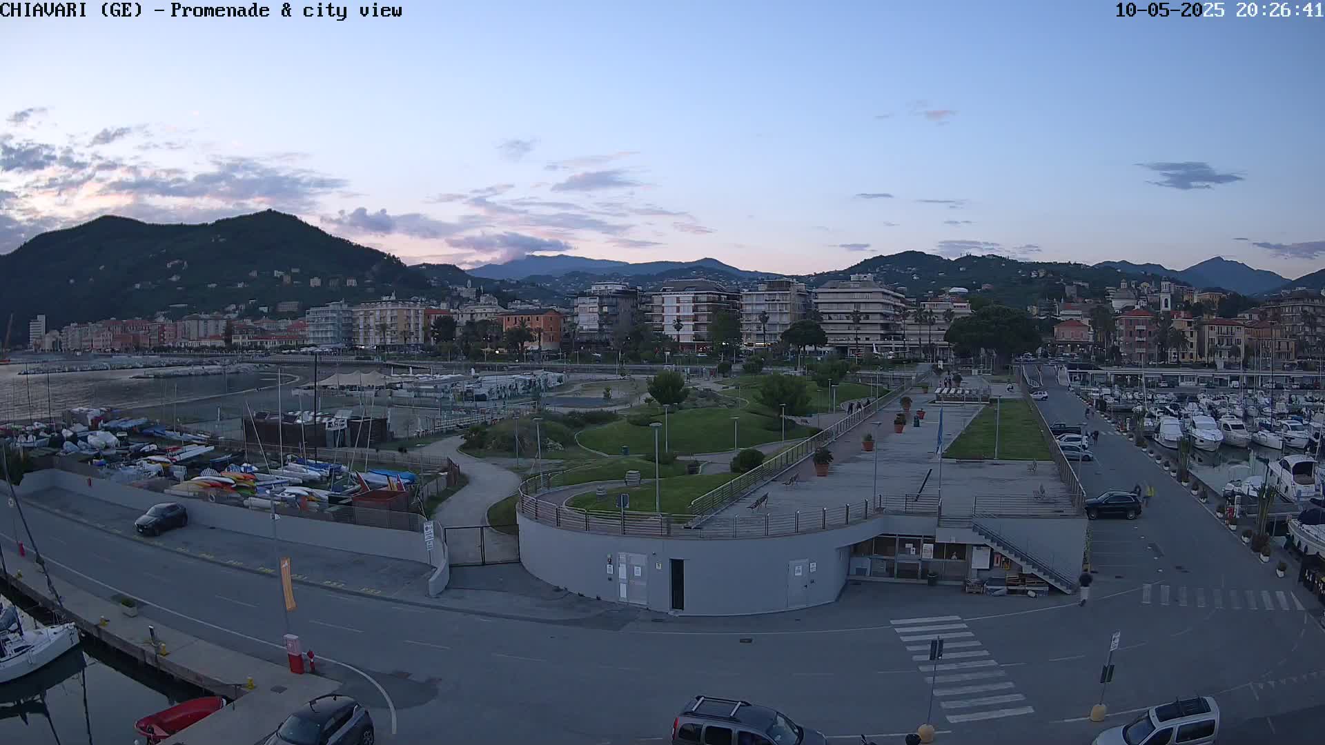 A seaside town at dusk, featuring a marina filled with boats, a promenade with a park, and apartment buildings against a backdrop of mountains under a mostly clear sky.