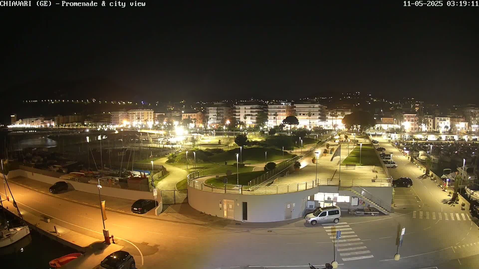 A nighttime aerial view of a harborside promenade and city, illuminated with artificial light, showing a marina, park, and buildings.