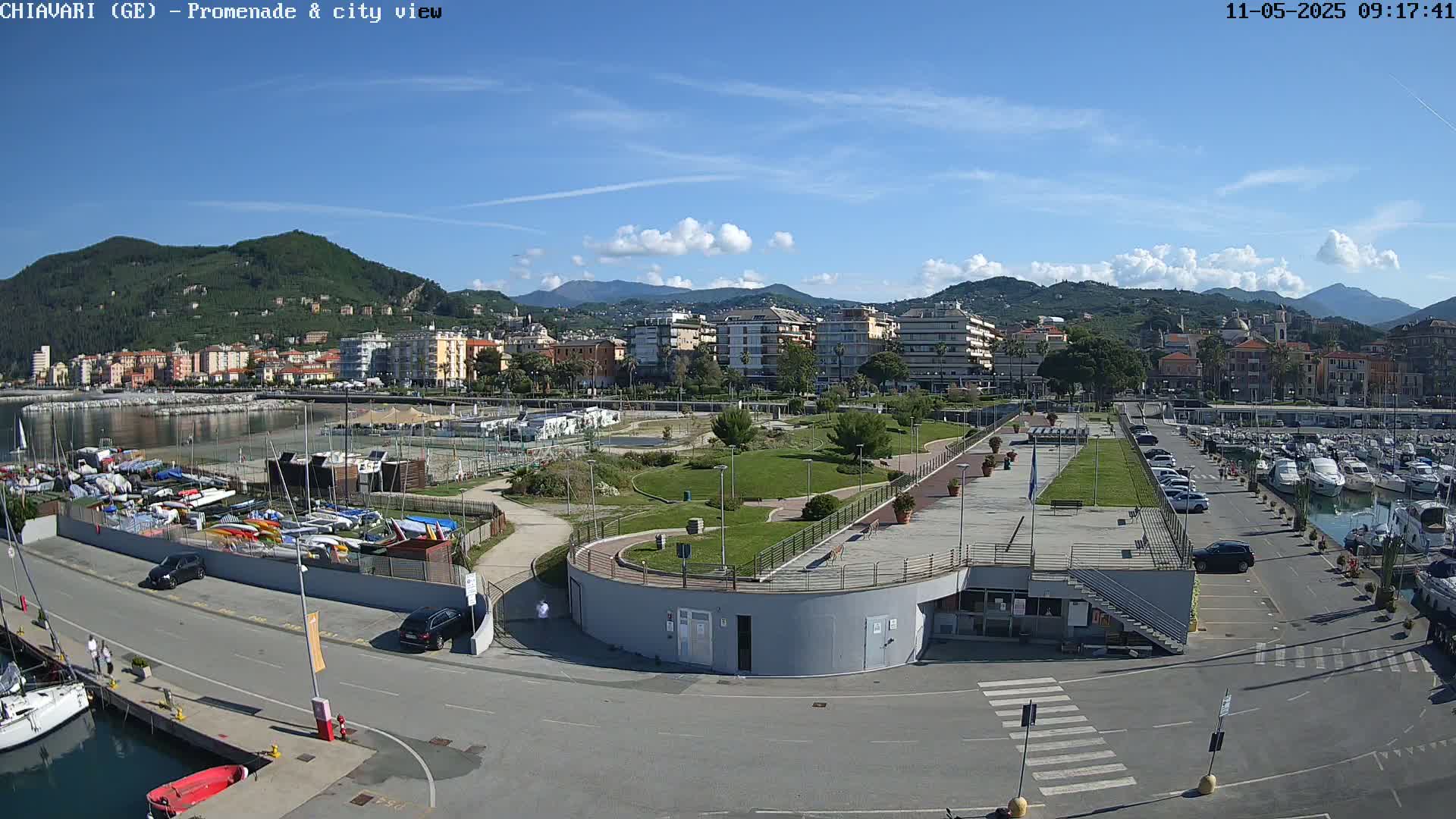 A sunny day reveals a harbor filled with boats, a seaside park with green lawns, and a town nestled against a hillside under a partly cloudy sky.