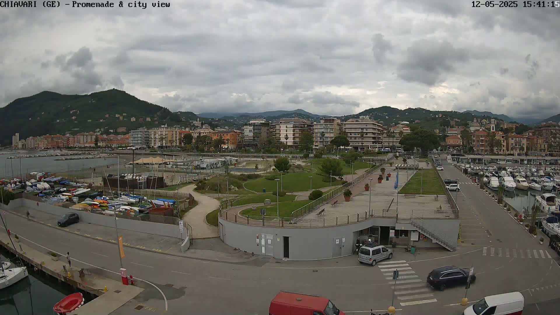 An overcast day reveals a waterfront promenade with a marina, a grassy park, and a cityscape nestled against hills in the background.