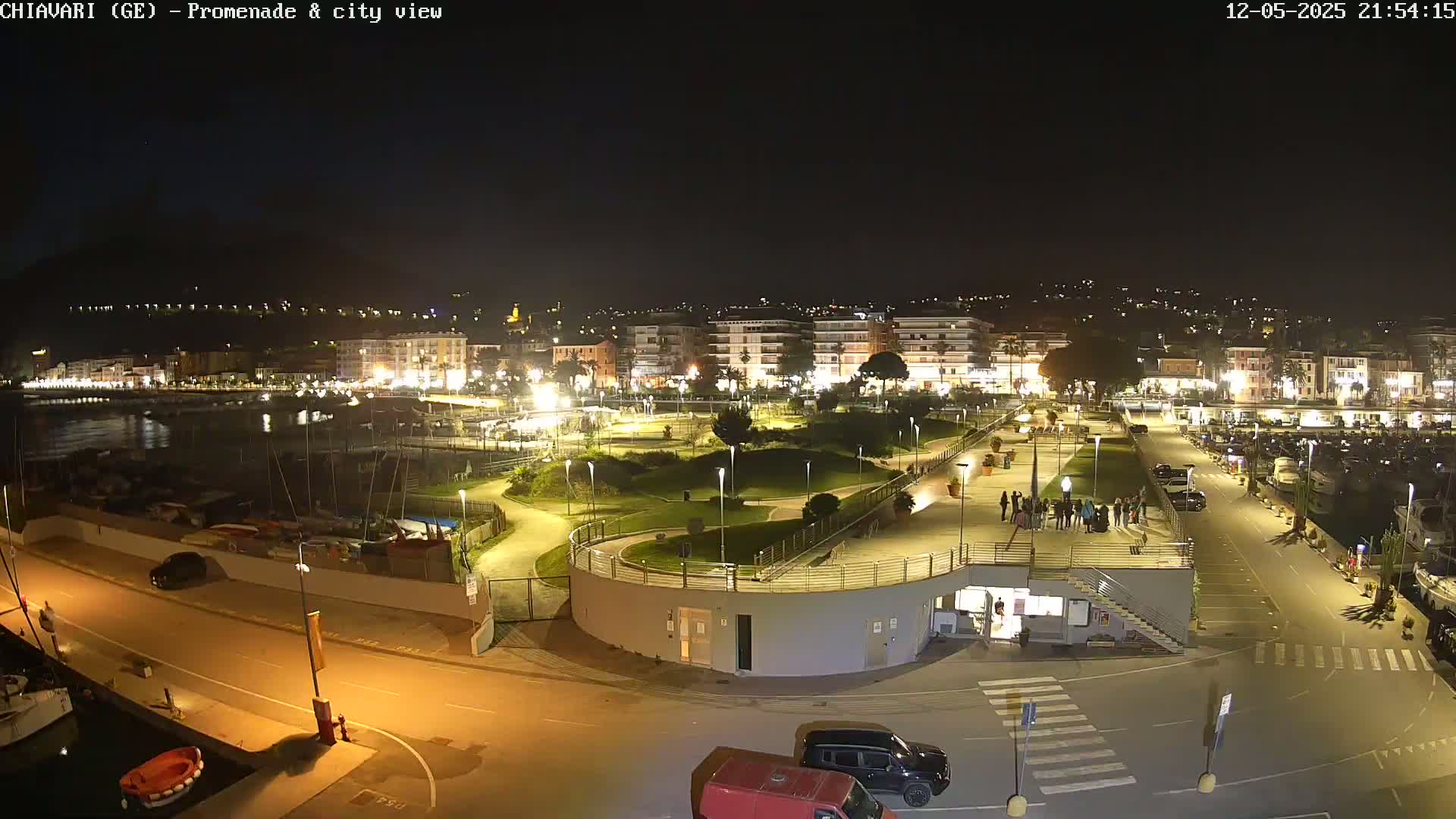 A nighttime, illuminated view of a waterfront promenade and city, featuring a marina, park, and roadways.