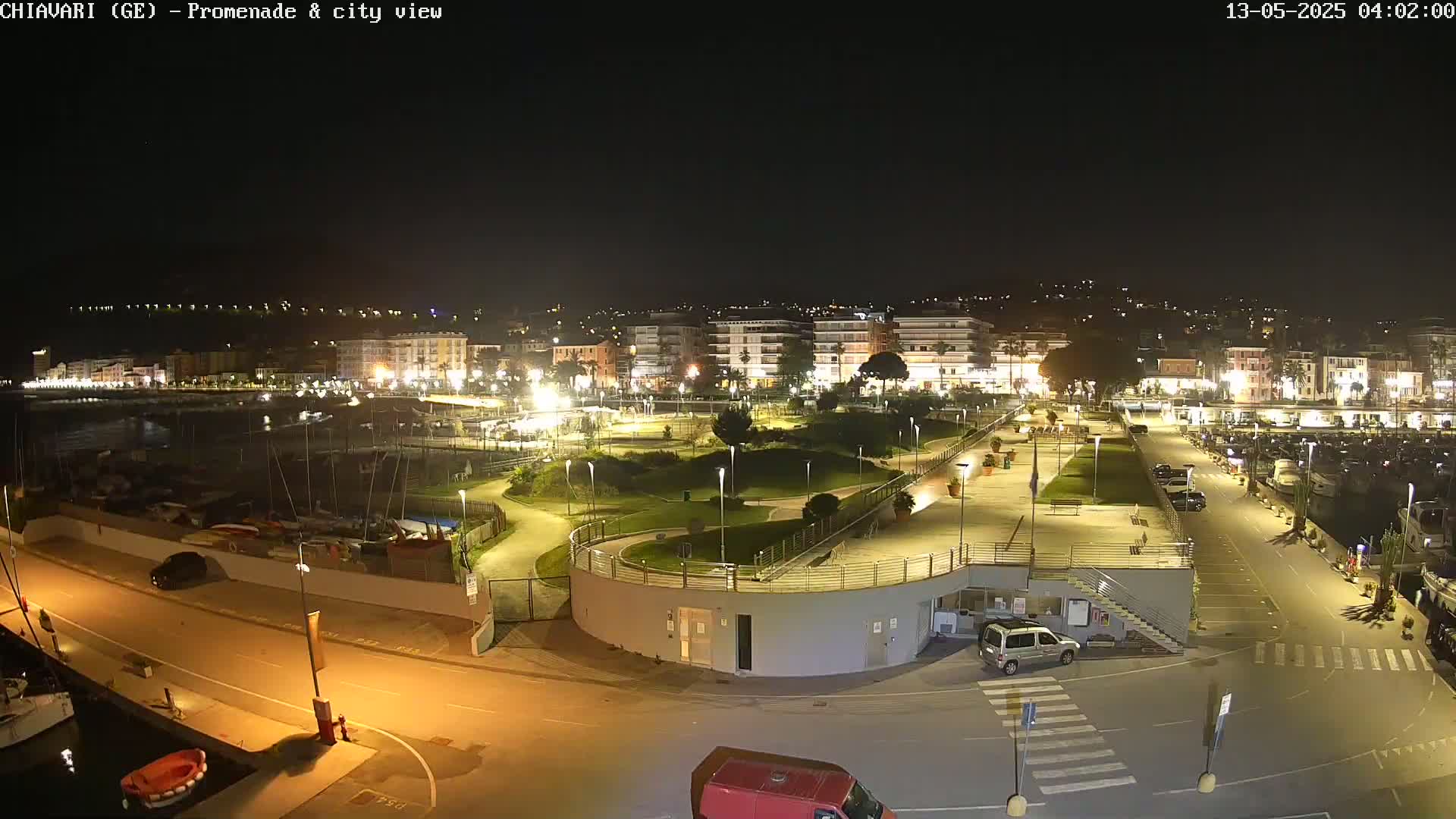 A nighttime aerial view of a harbor, promenade, and city, with a small park and parking lot visible.