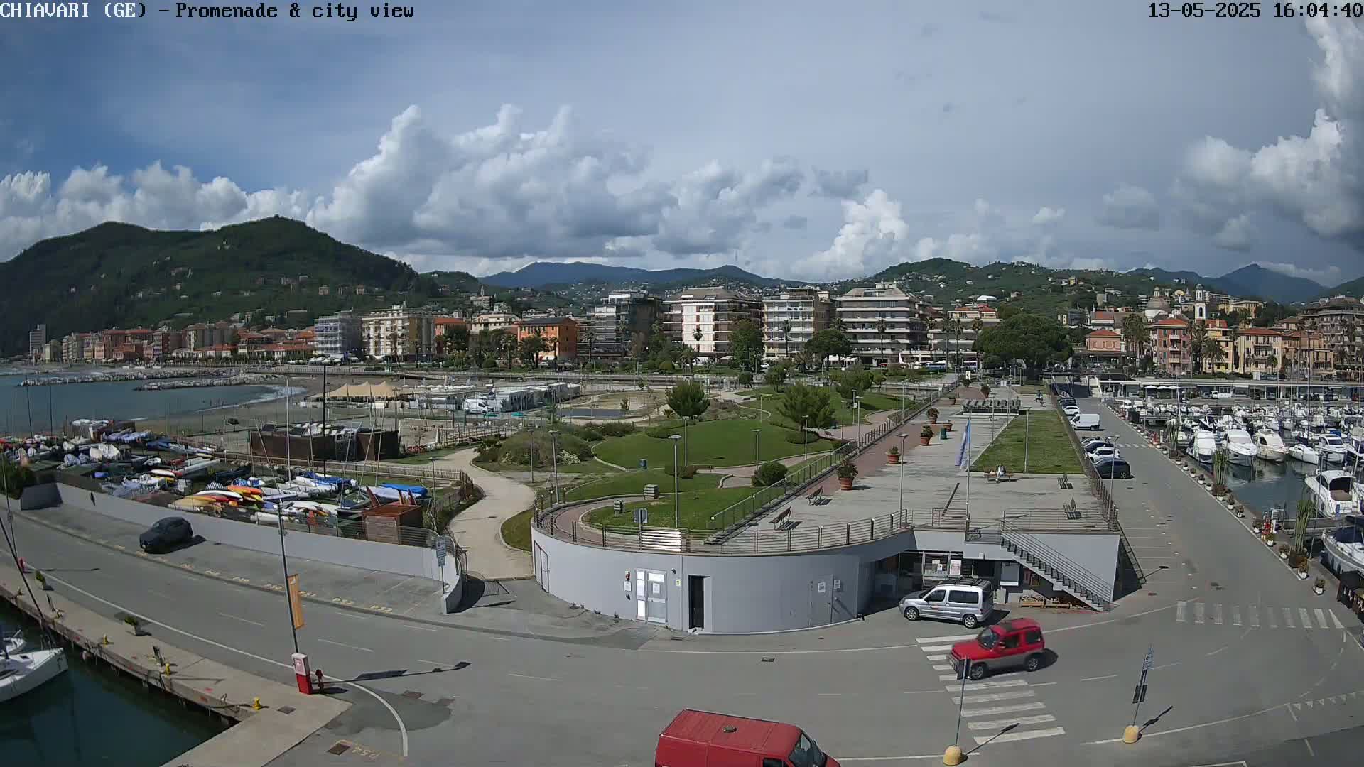 A coastal city scene on a partly cloudy day shows a harbor filled with boats, a promenade with landscaped gardens, and buildings nestled against a hillside.
