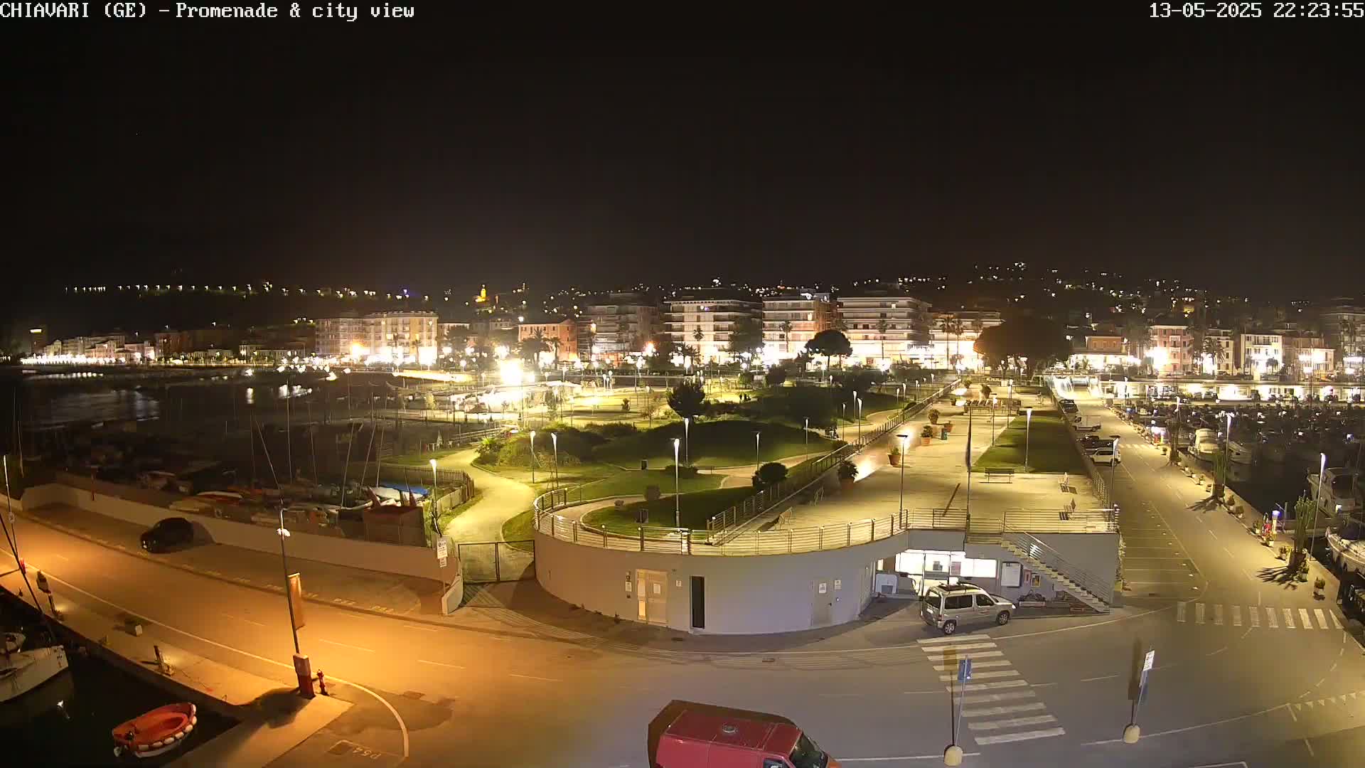A nighttime view of a city's waterfront promenade, showing a harbor filled with boats, a park-like area with pathways, and buildings lit up along the shoreline.