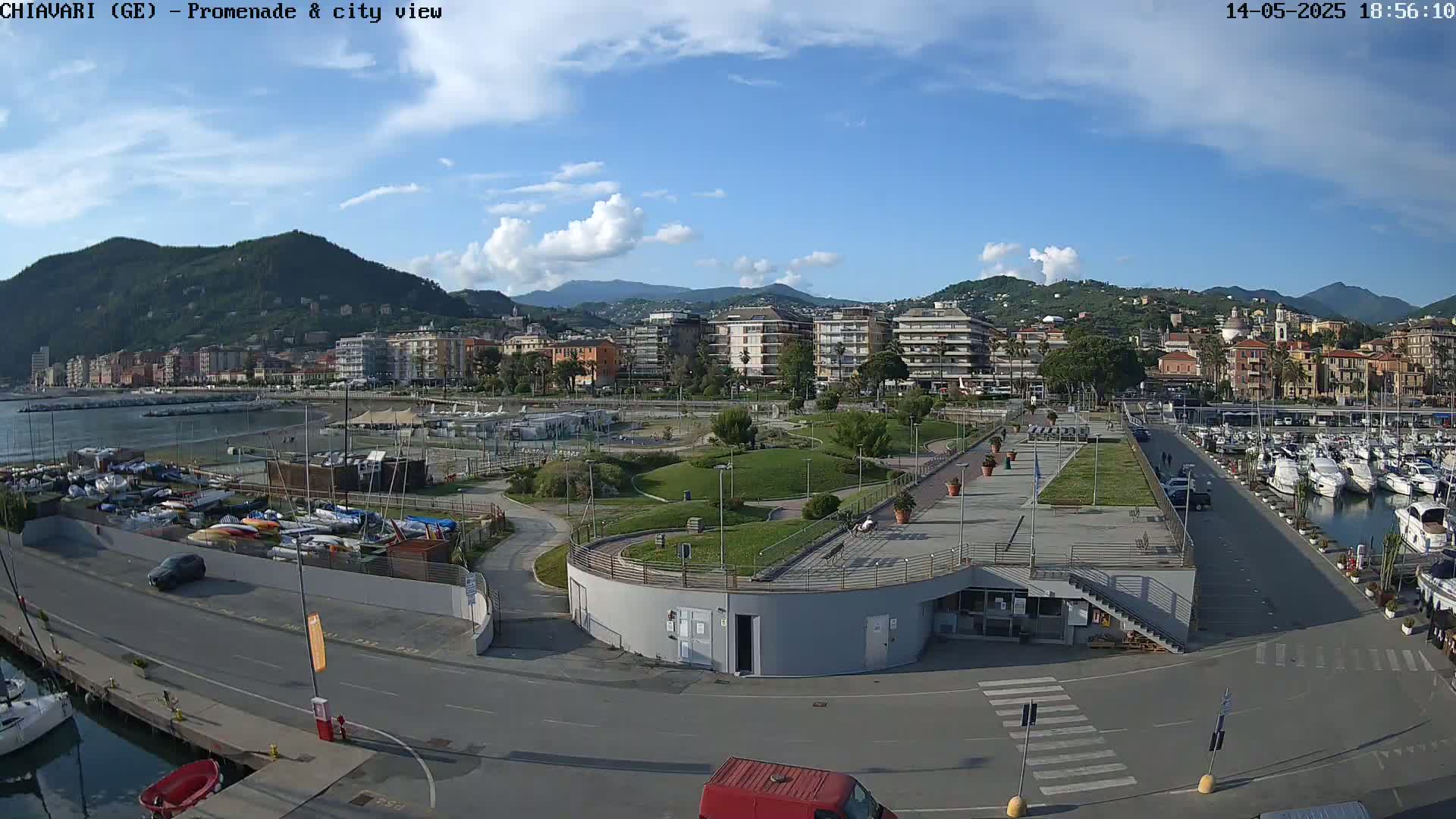 A partly sunny day overlooking a coastal city with a marina, promenade, and park-like area, backed by mountains.