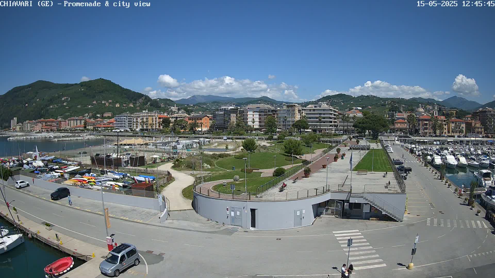 An aerial view of a coastal city on a sunny day shows a harbor filled with boats, a park with paved walkways, and buildings nestled against hills in the background.