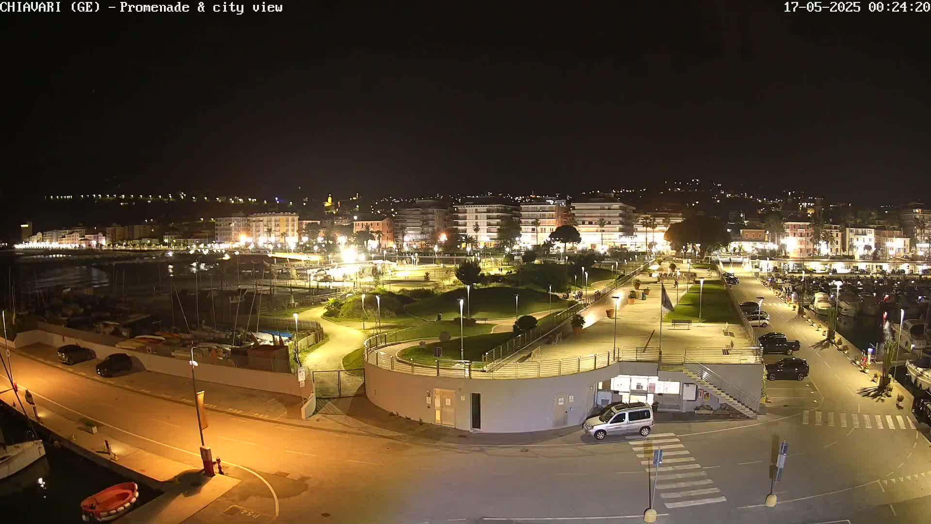 A nighttime, brightly lit view of a harbor, promenade, and city with a few cars parked in lots.