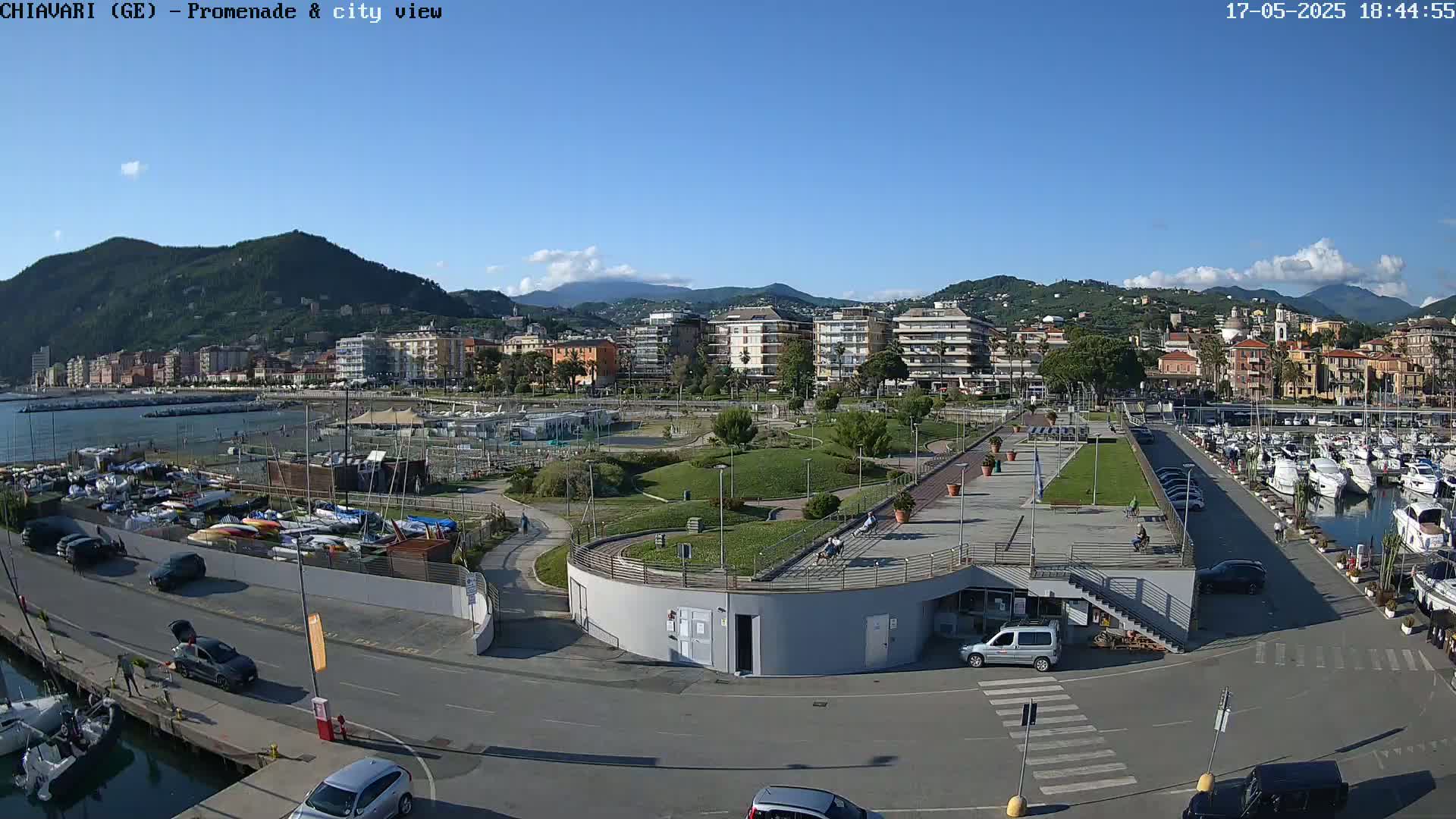 An aerial view of a waterfront promenade and marina on a sunny day, with a town and mountains in the background.