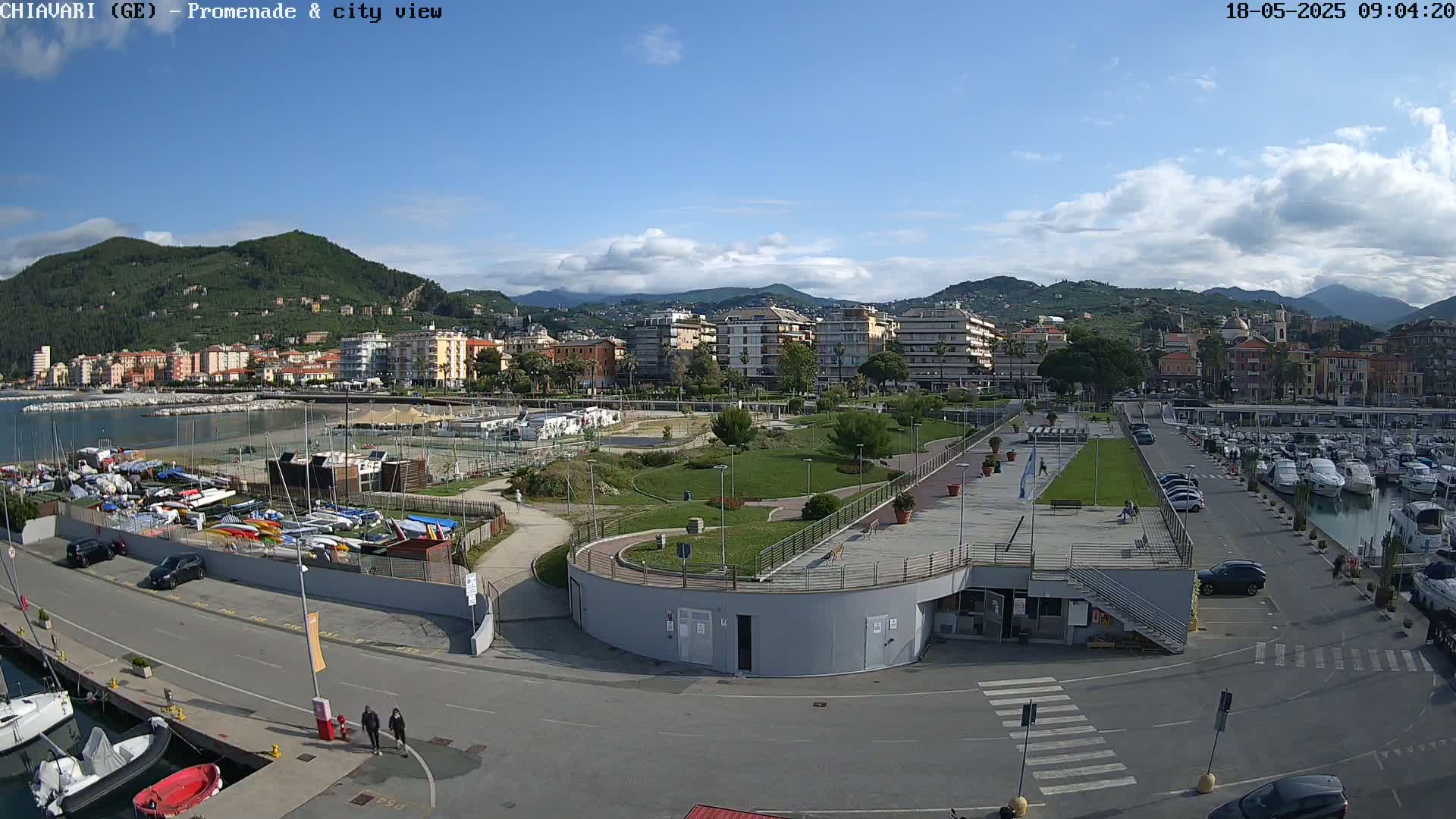 A coastal city scene under partly cloudy skies, showing a marina filled with boats, a park with pathways, and buildings stretching along the shoreline with hills in the background.