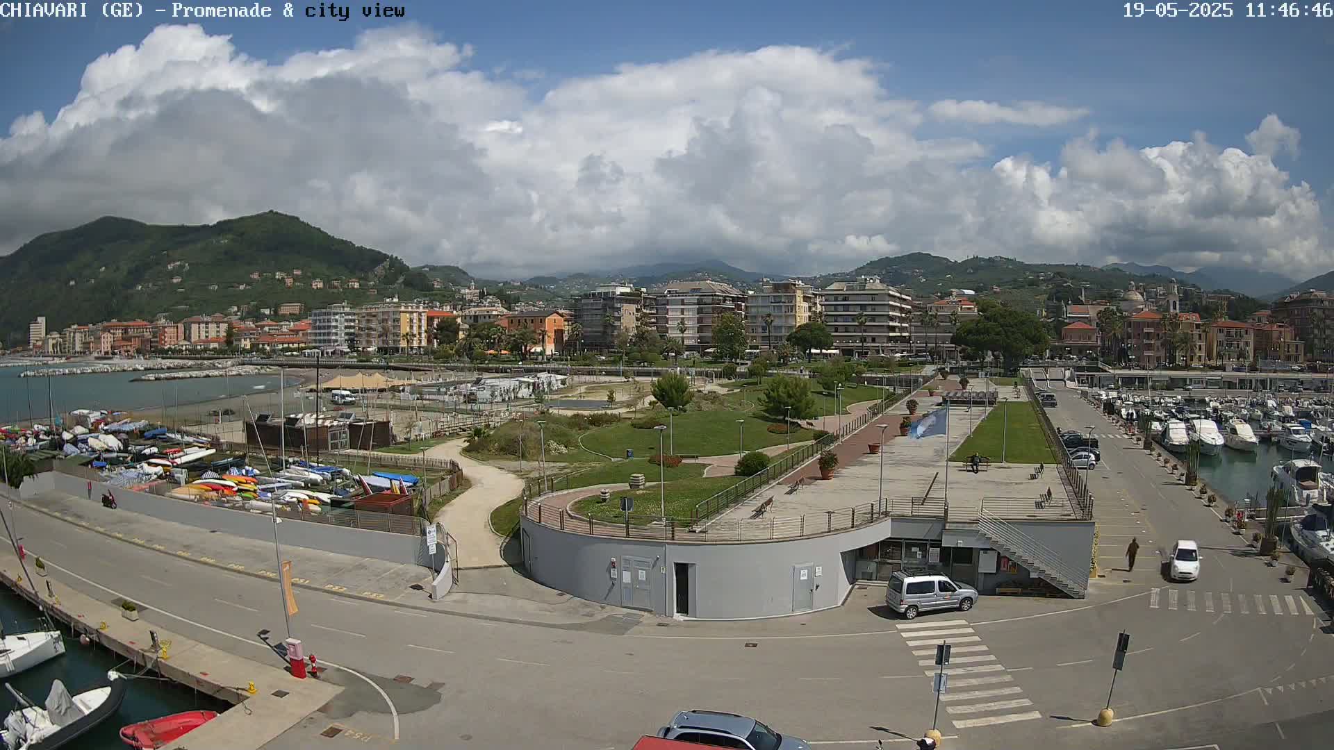 A partly cloudy day reveals a coastal town with a marina, a promenade, and a park next to a waterfront with many boats and buildings stretching into the hills in the background.