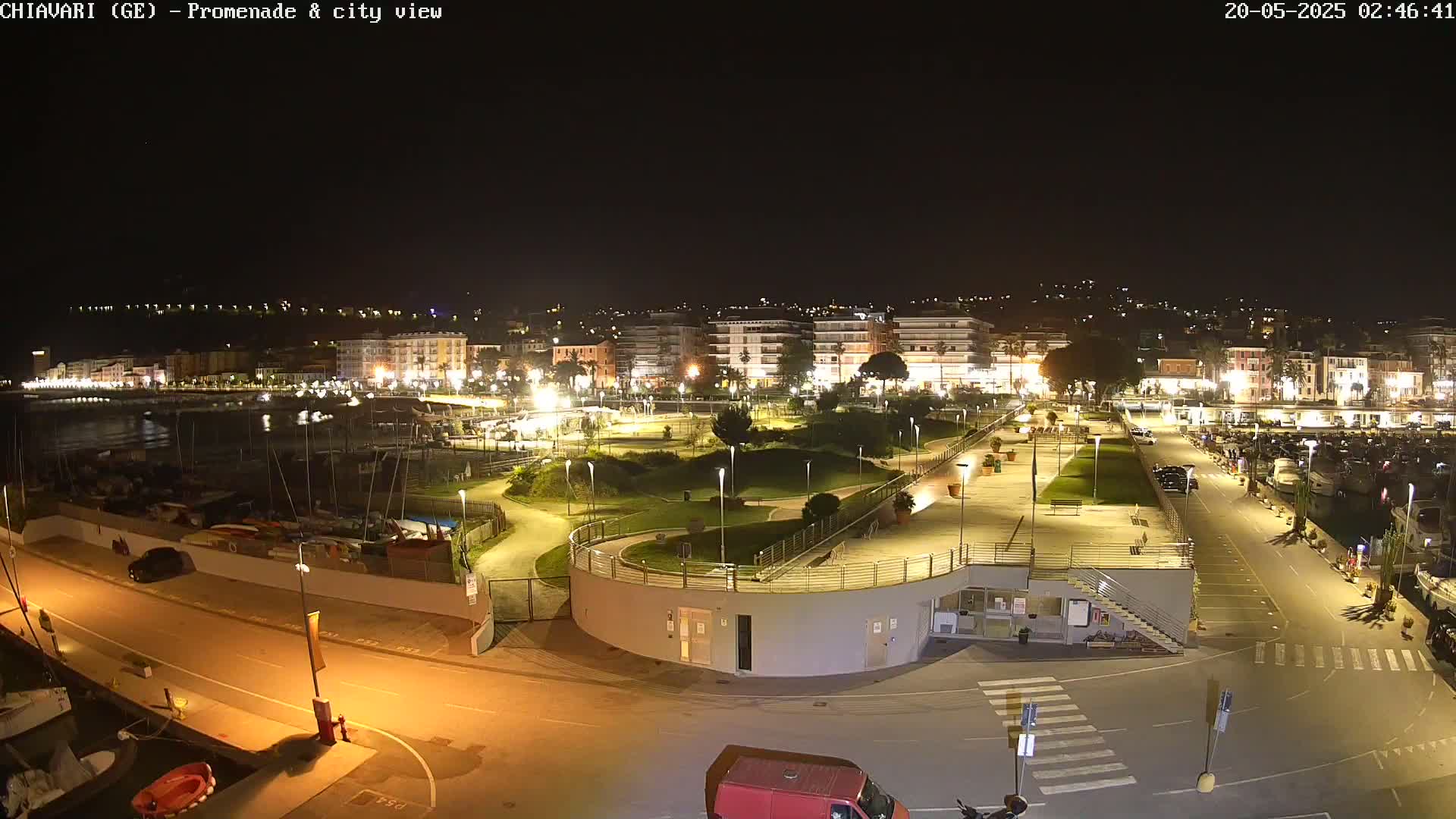 A nighttime aerial view of a harbor town's promenade and marina, illuminated by streetlights, showing boats in the water and buildings along the shoreline.