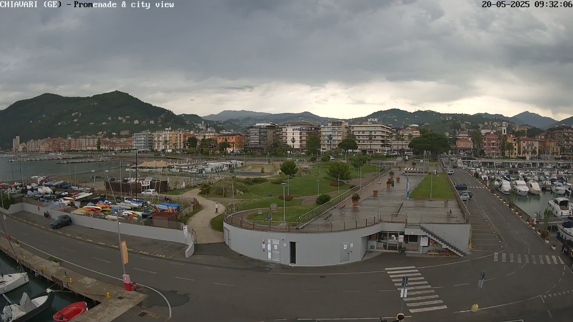 A waterfront promenade and city, under a cloudy sky, features a marina filled with boats, a grassy park, and buildings extending along a coastline backed by hills.