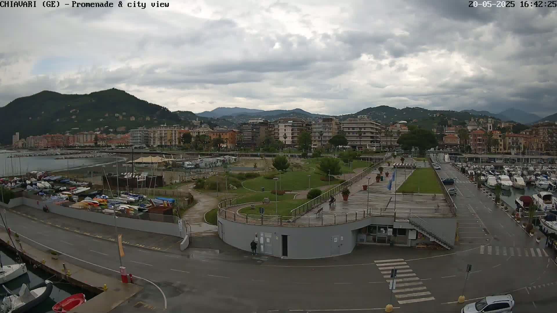 A coastal city with a harbor full of boats, a promenade with a park, and mountains in the background under a cloudy sky.
