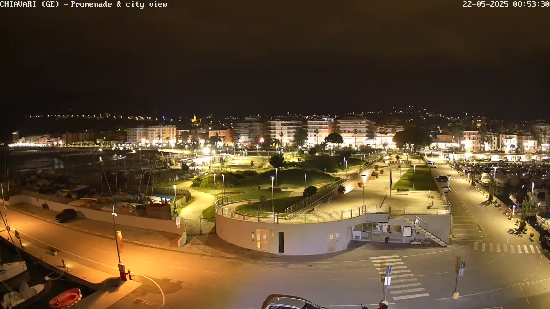 A nighttime aerial view of a city's waterfront promenade and marina, brightly lit with streetlights, showing a harbor with boats and cars parked nearby.