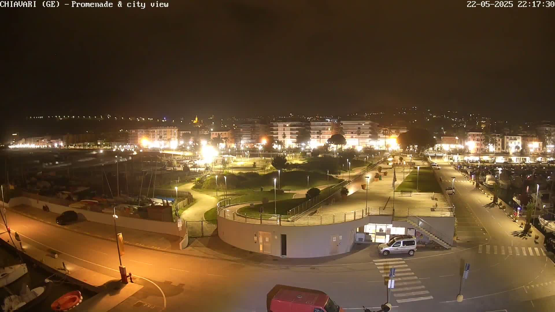 A nighttime aerial view of a city's illuminated waterfront promenade and marina, featuring a park, buildings, and parked cars.