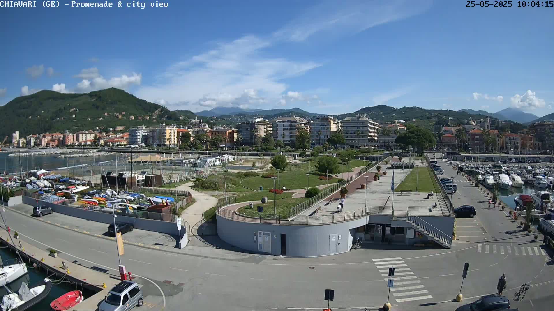 A waterfront promenade and marina are shown on a sunny day, with a town and mountains in the background.