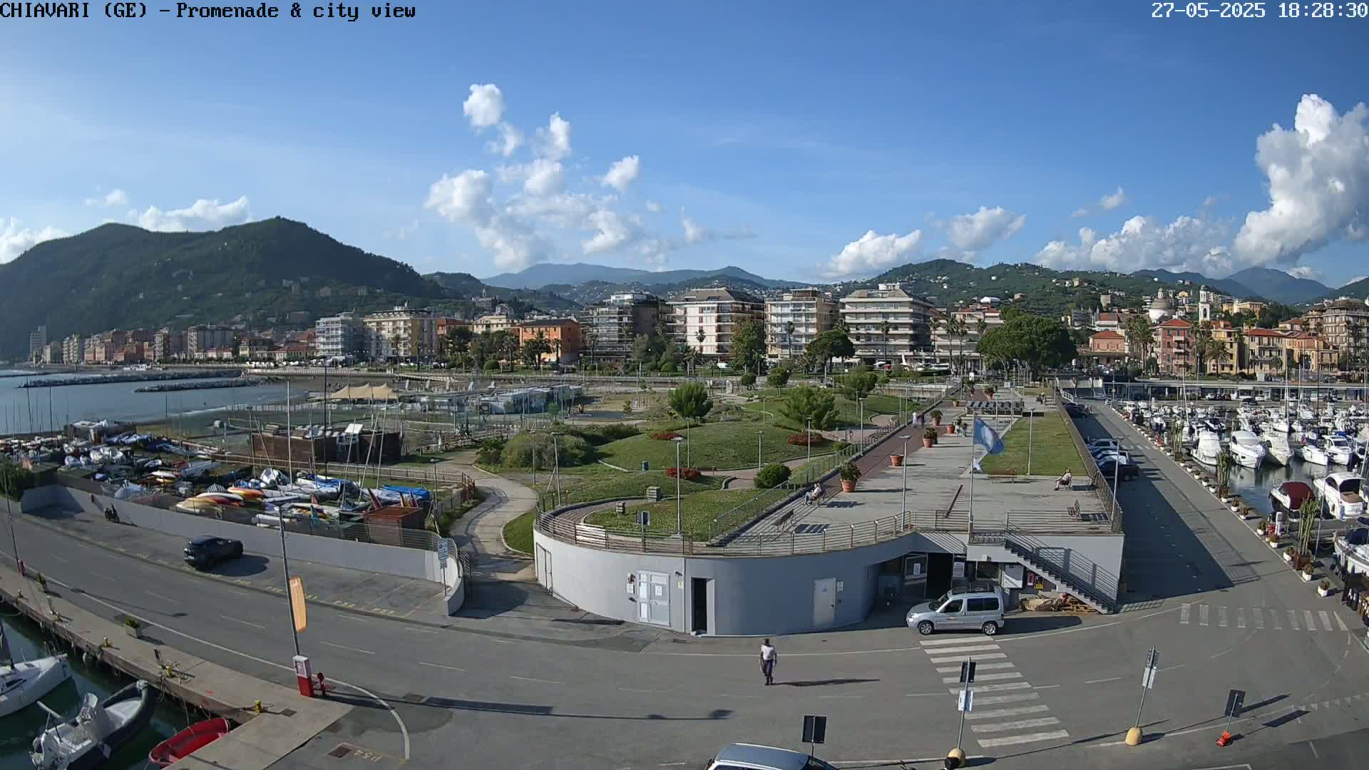 An aerial view shows a harbor with numerous boats, a paved promenade lined with parked cars, a landscaped park, and a city with buildings nestled against mountains under a partly cloudy sky.