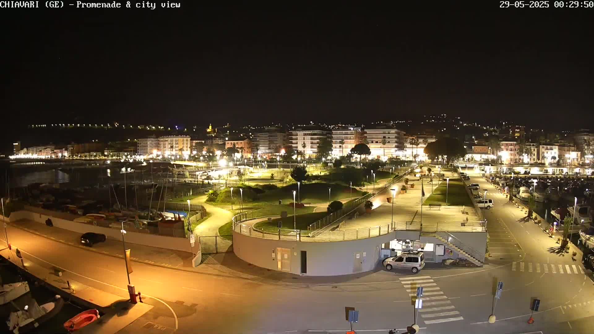 A nighttime view of a city's illuminated harbor and promenade, featuring a marina, park, and buildings along the waterfront.