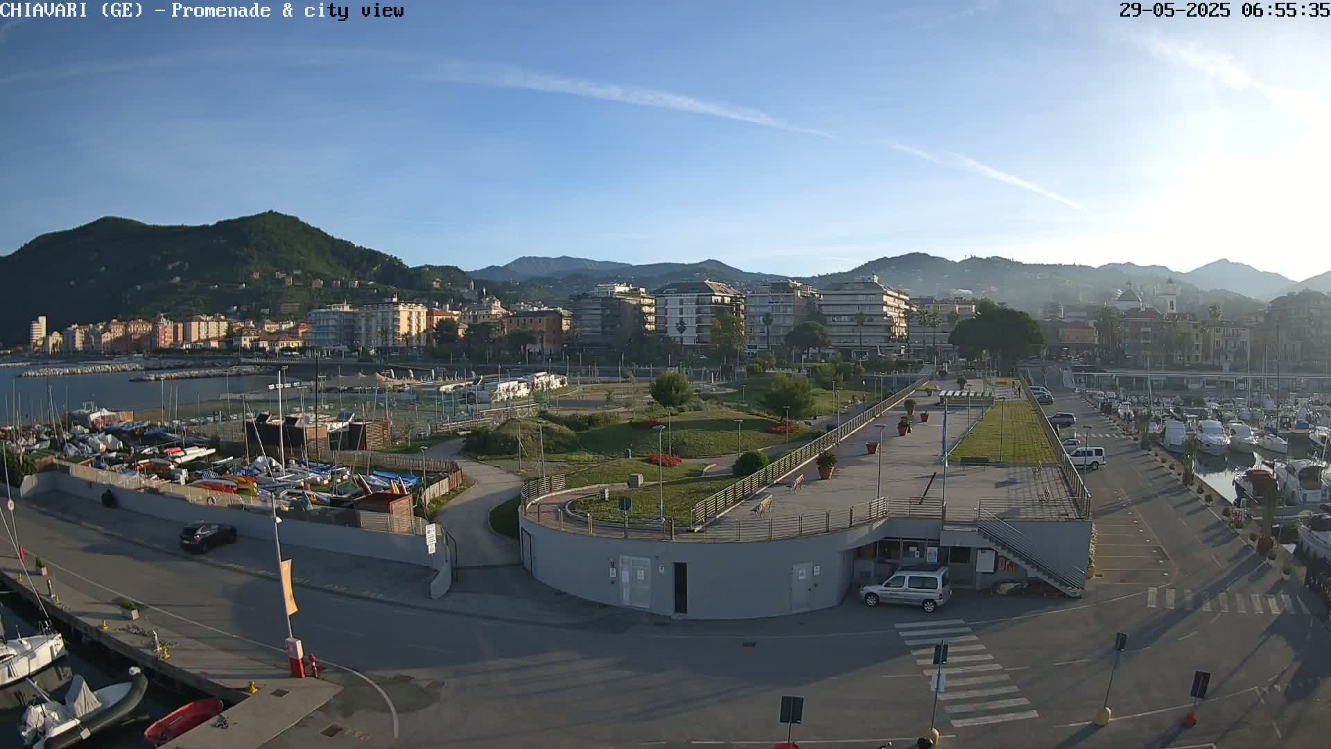 A seaside town is visible under a clear blue sky, with a marina filled with boats and a promenade overlooking a park and city buildings nestled against hills.