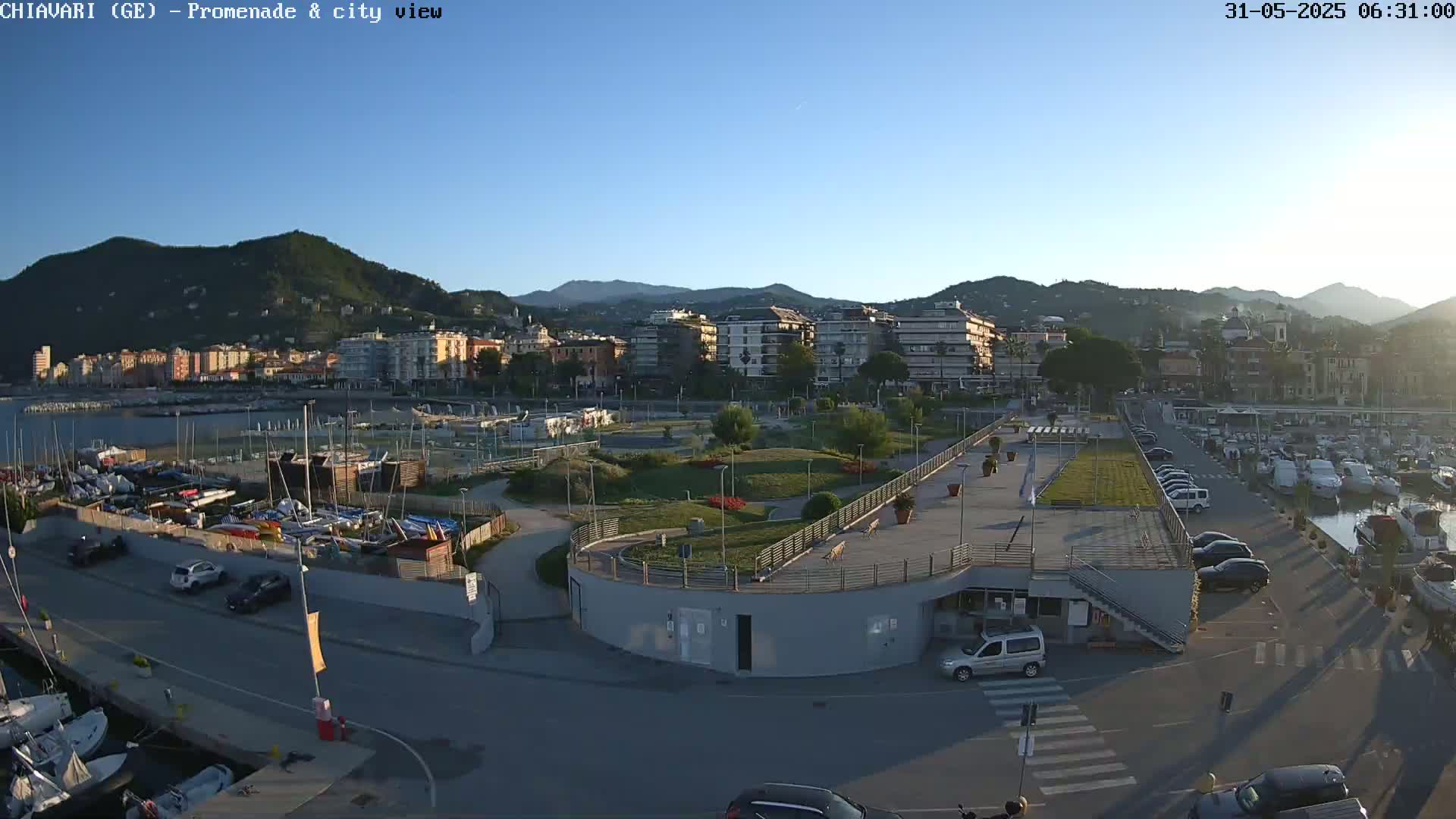 A coastal city scene under a clear, sunny sky shows a marina filled with boats, a promenade with cars parked along it, and a park with green landscaping separating the waterfront from a line of buildings nestled against hills.