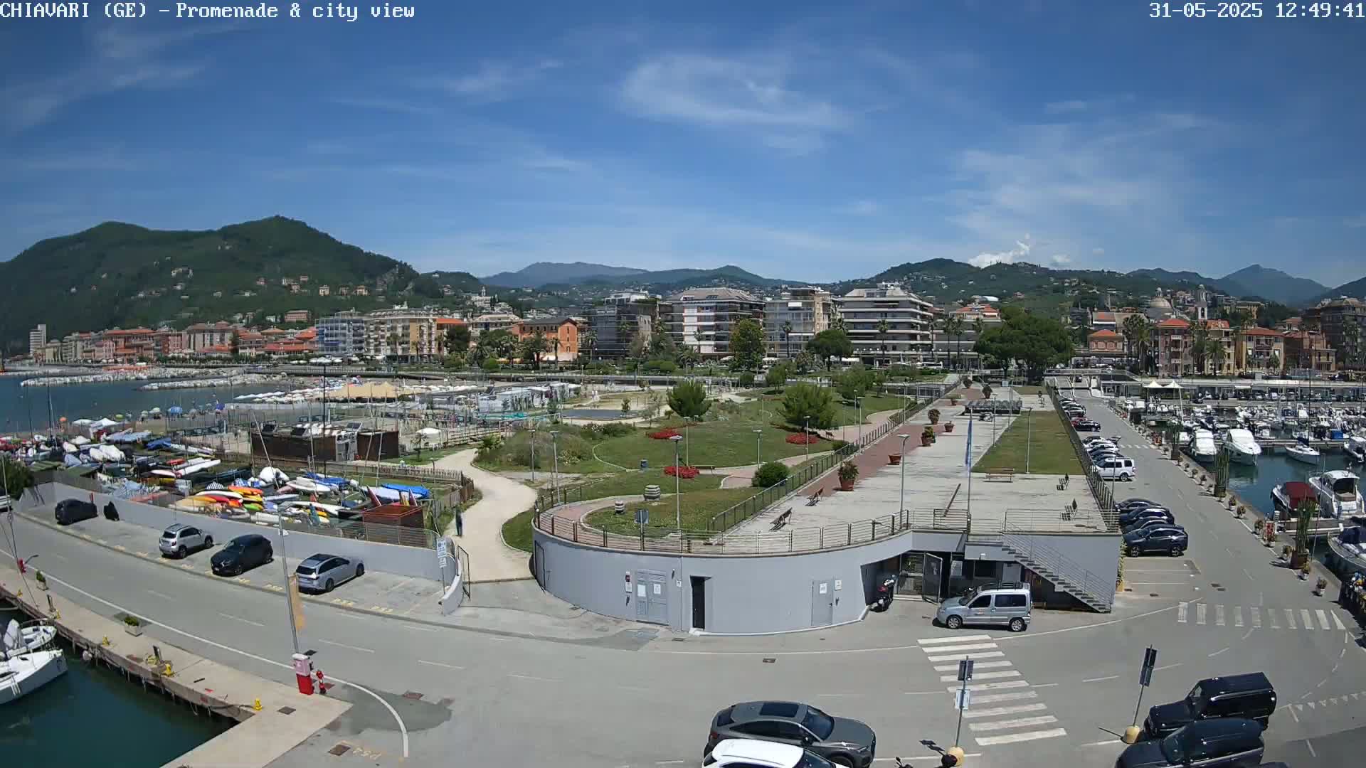 A seaside town nestled against mountains under a clear blue sky, featuring a marina filled with boats, a park, and a parking lot with cars.