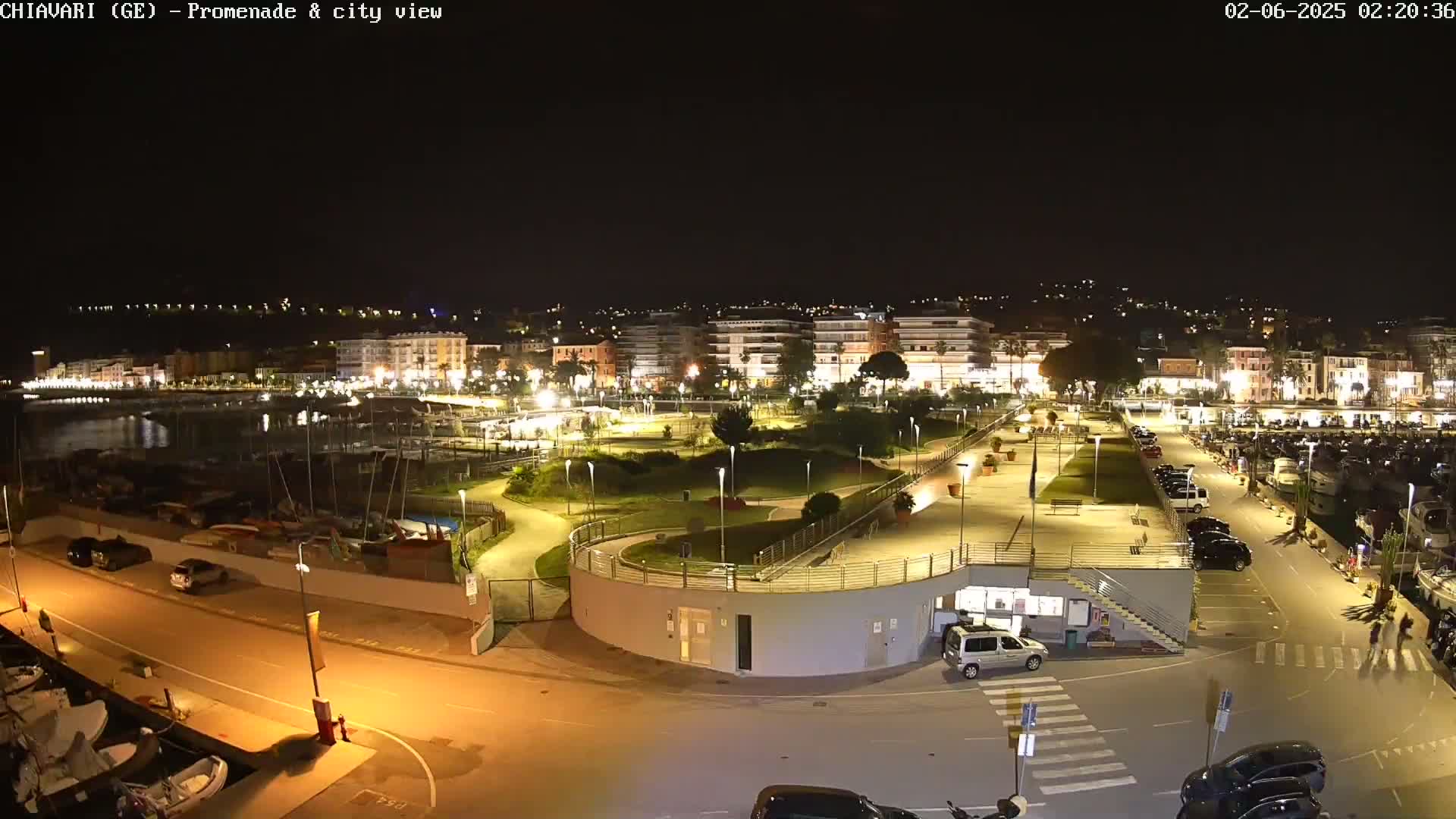 A nighttime aerial view of a waterfront promenade and city, with boats in a marina, parking lots, and a park illuminated with lights.