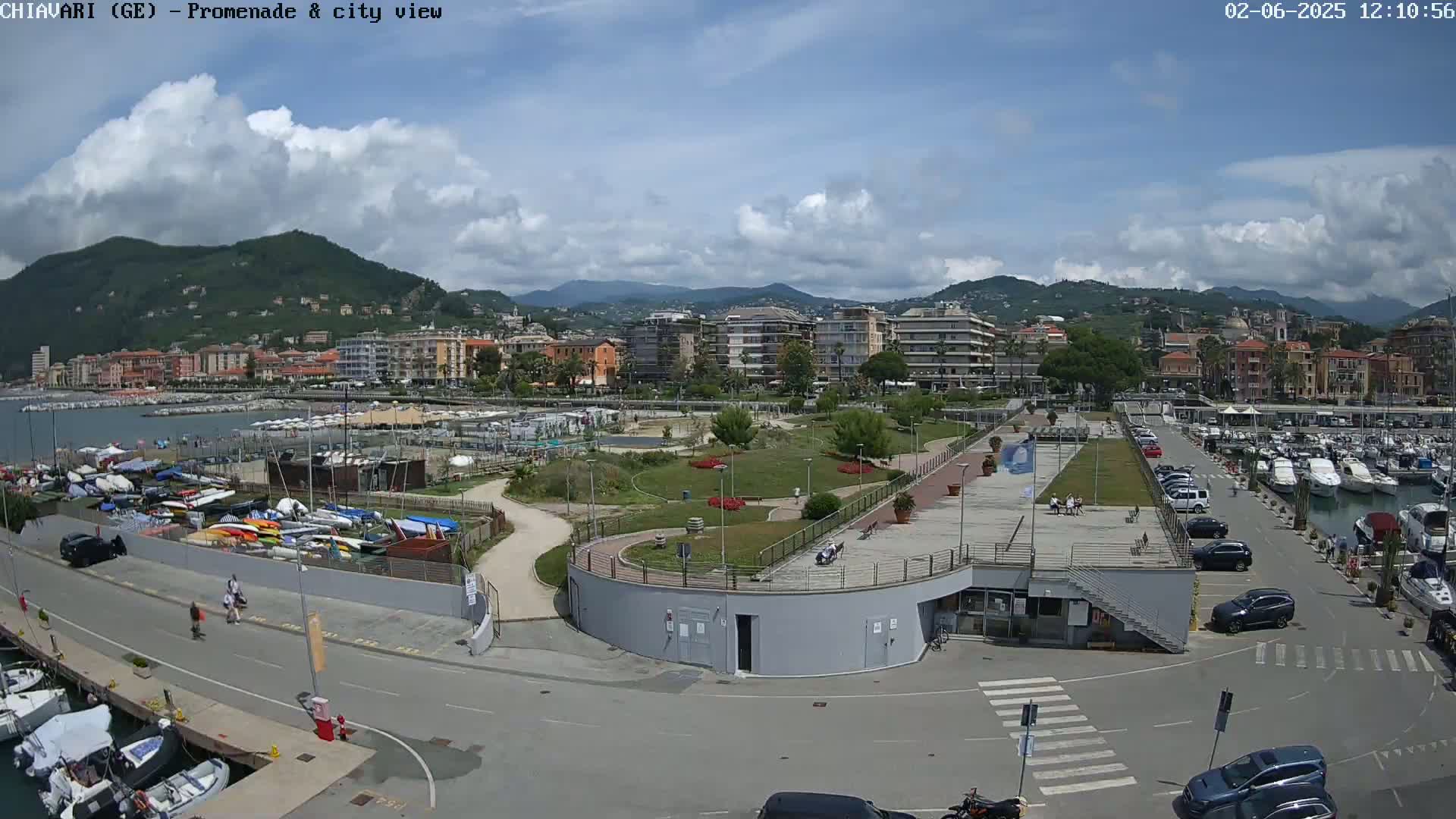 A partly cloudy day reveals a coastal city scene with a marina filled with boats, a promenade, and a park, all situated against a backdrop of mountains.