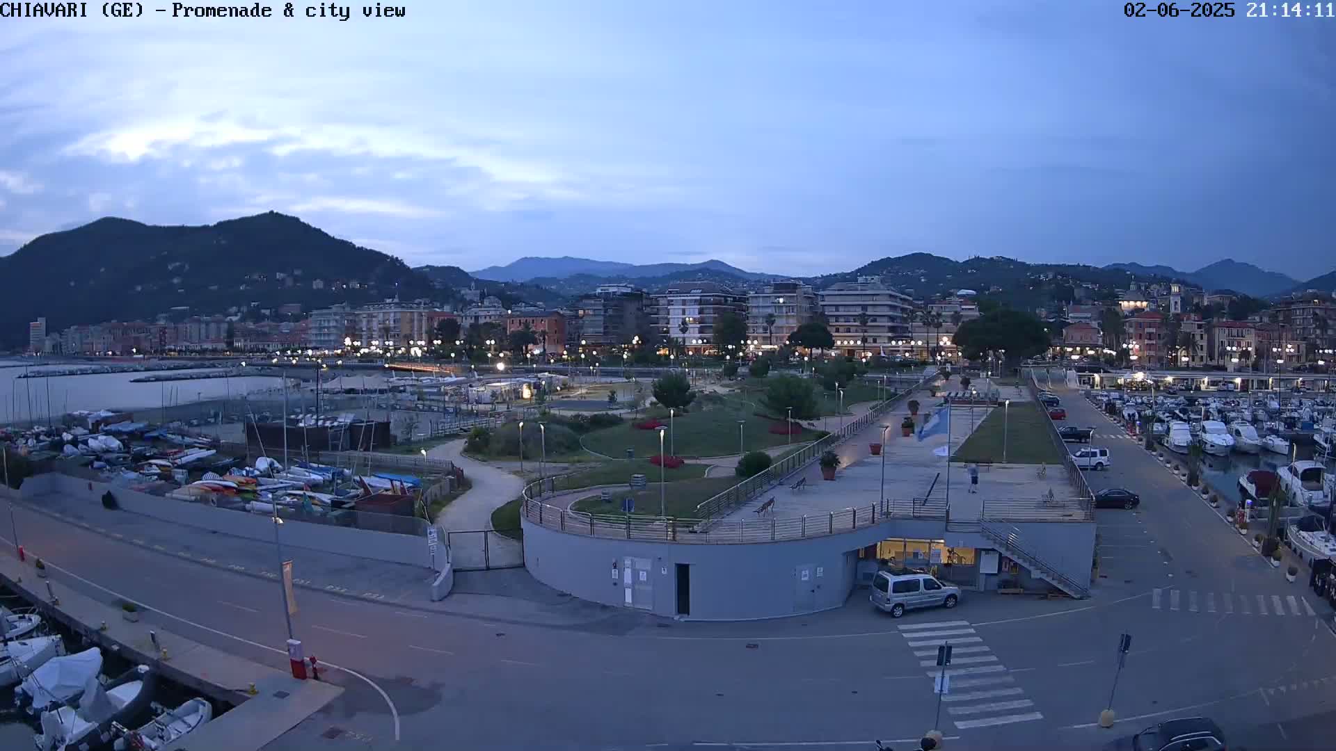 An aerial view of a coastal town at dusk, showing a harbor filled with boats, a promenade with a park, and buildings against a backdrop of mountains under a clear sky.