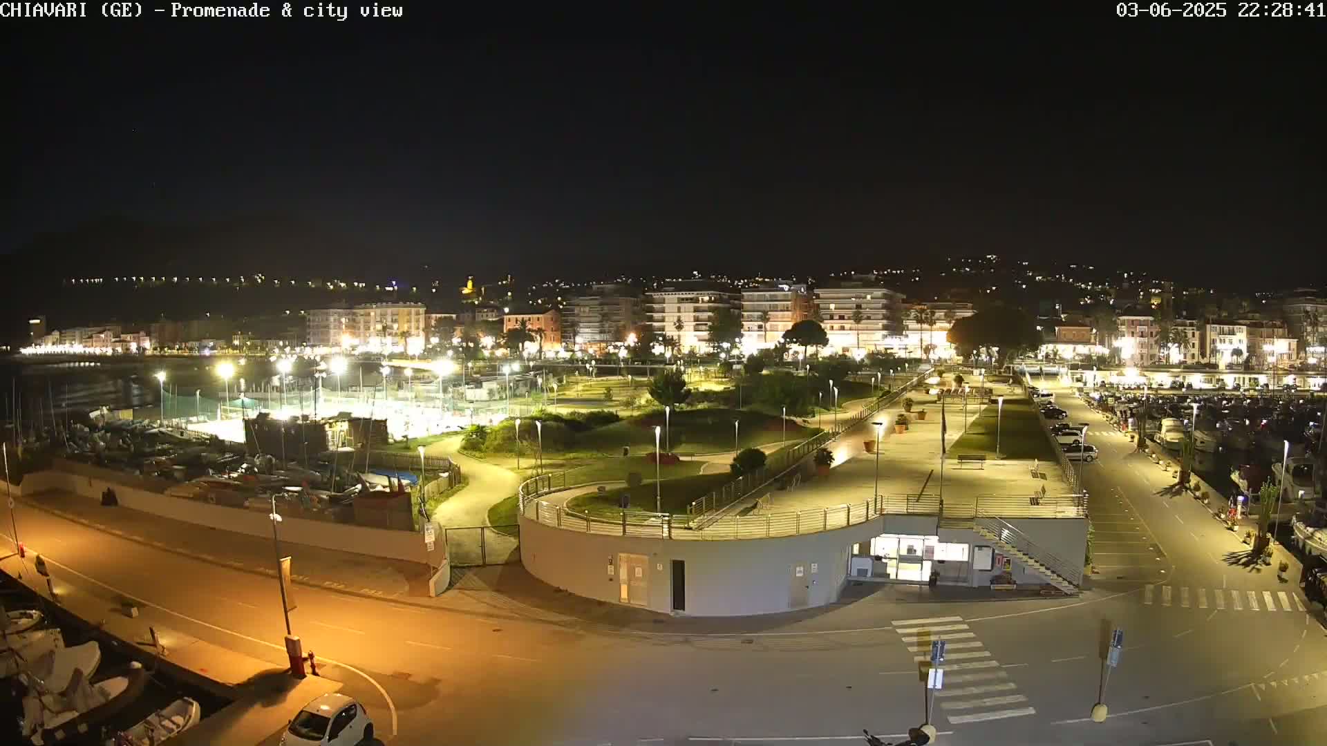 A nighttime view of a city's waterfront promenade, featuring a park, parking lot, and harbor filled with boats, all brightly lit under a clear, dark sky.