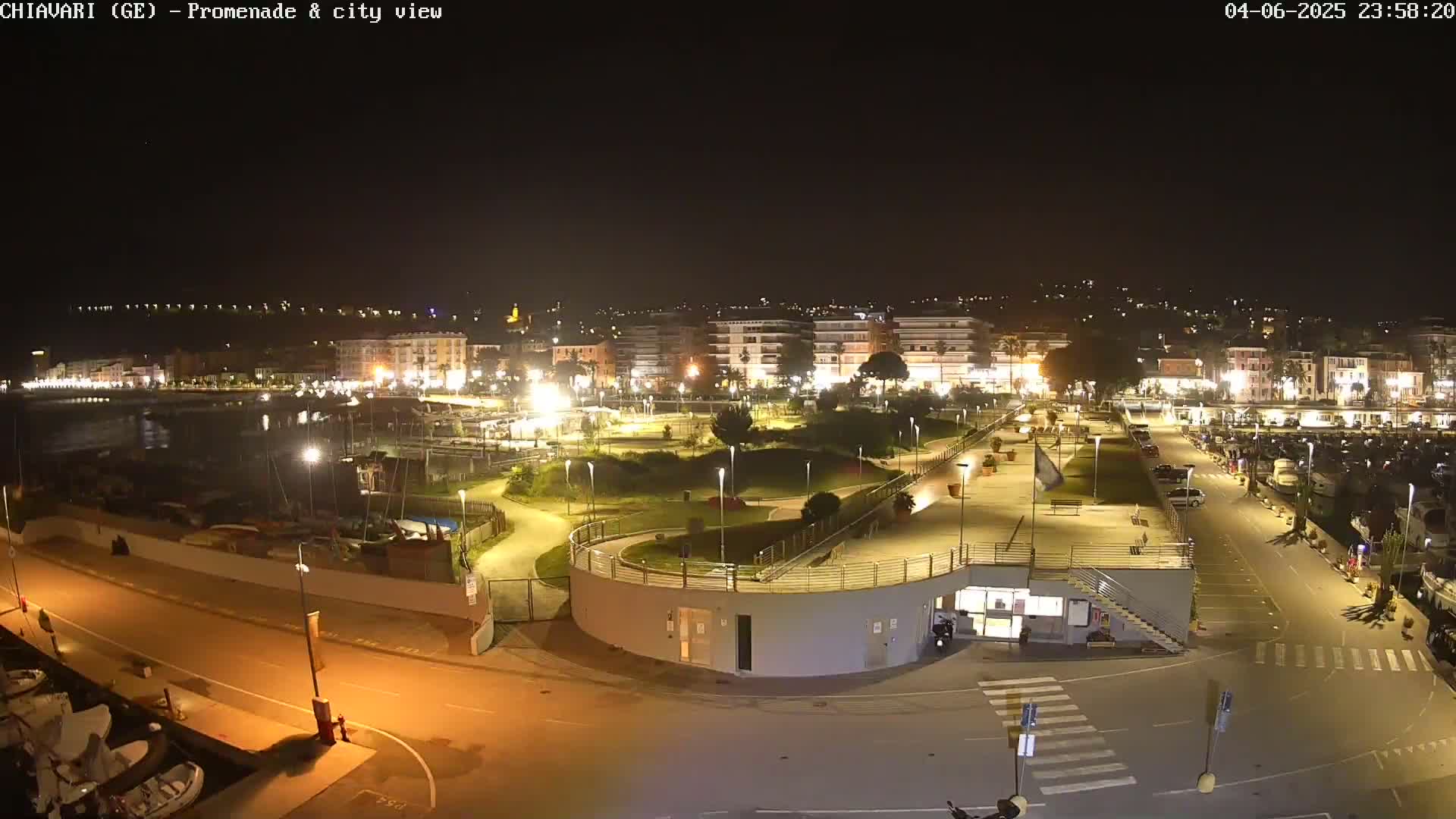 A nighttime aerial view of a brightly lit waterfront promenade and city, featuring a marina with boats, a park with winding paths, and a parking lot.