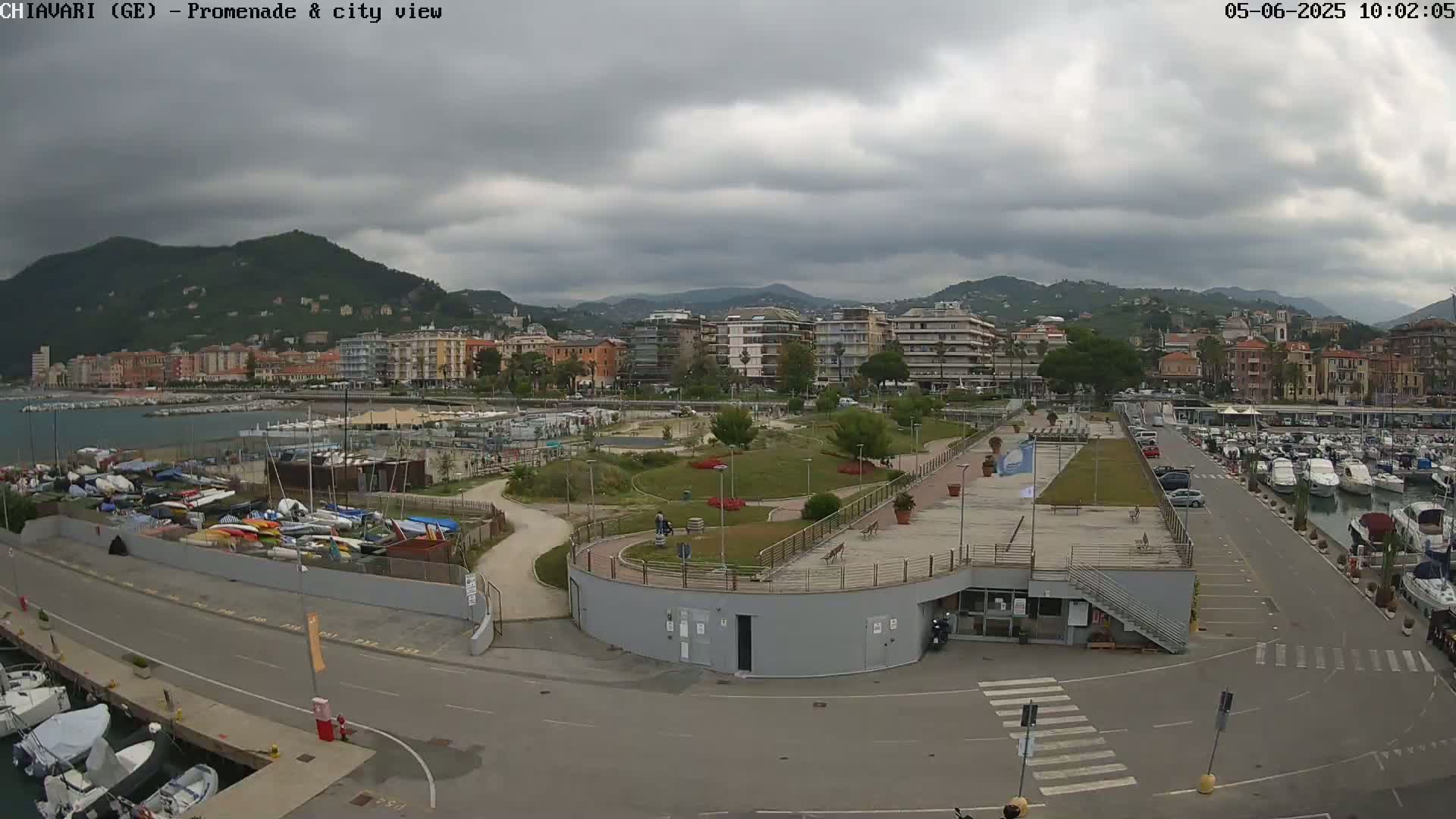 An aerial view shows a harbor filled with boats next to a paved promenade and a grassy park, all overlooking a city nestled against hills under an overcast sky.