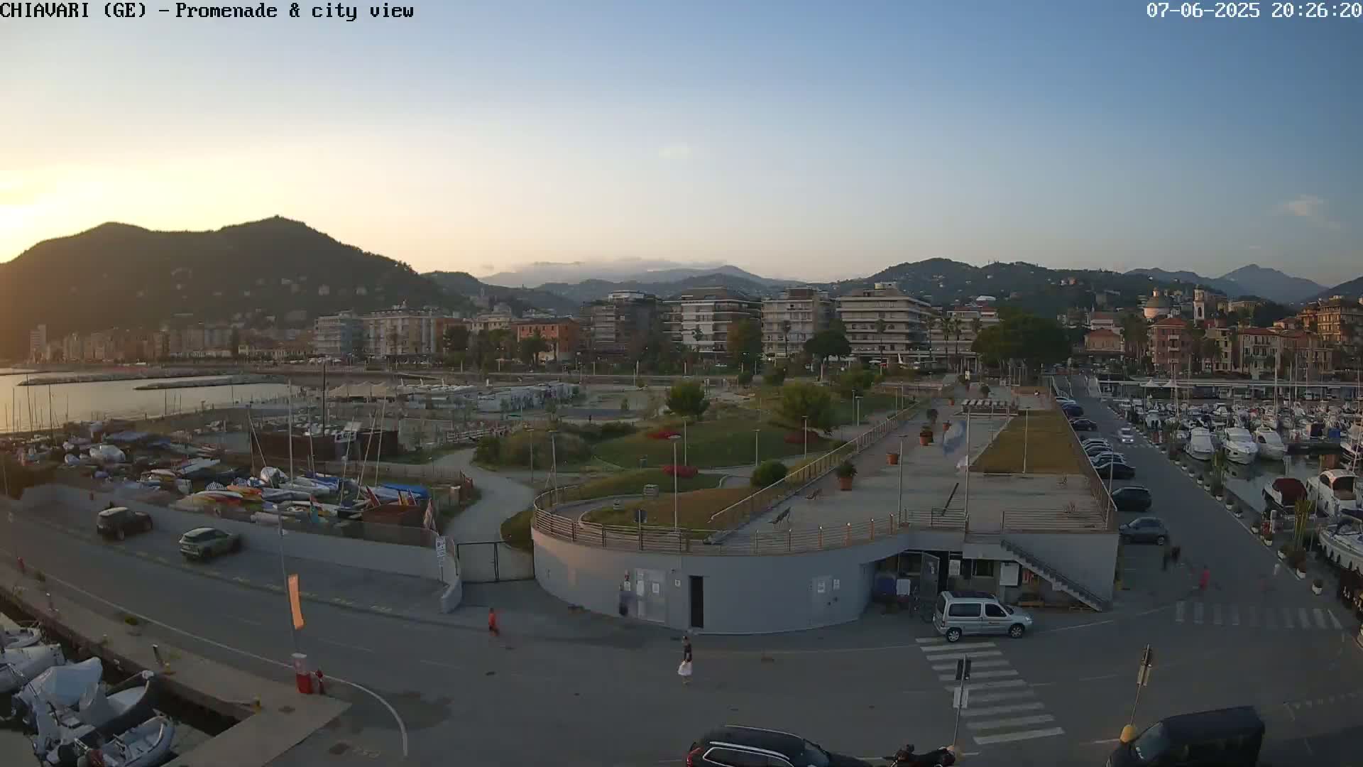An aerial view shows a seaside town at dusk, with a marina full of boats and cars parked along a promenade next to a park and a curving building.