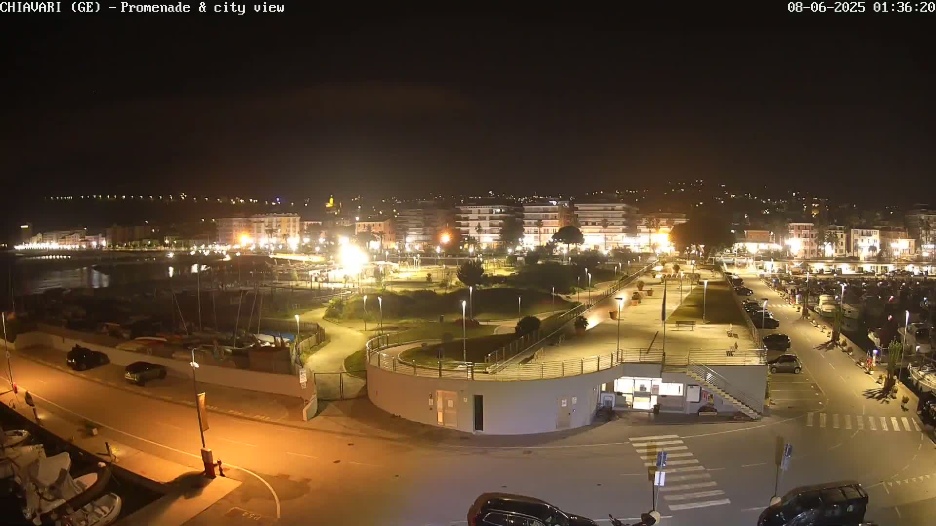 A nighttime view of a city's waterfront promenade, harbor, and parking areas, illuminated by artificial lights.