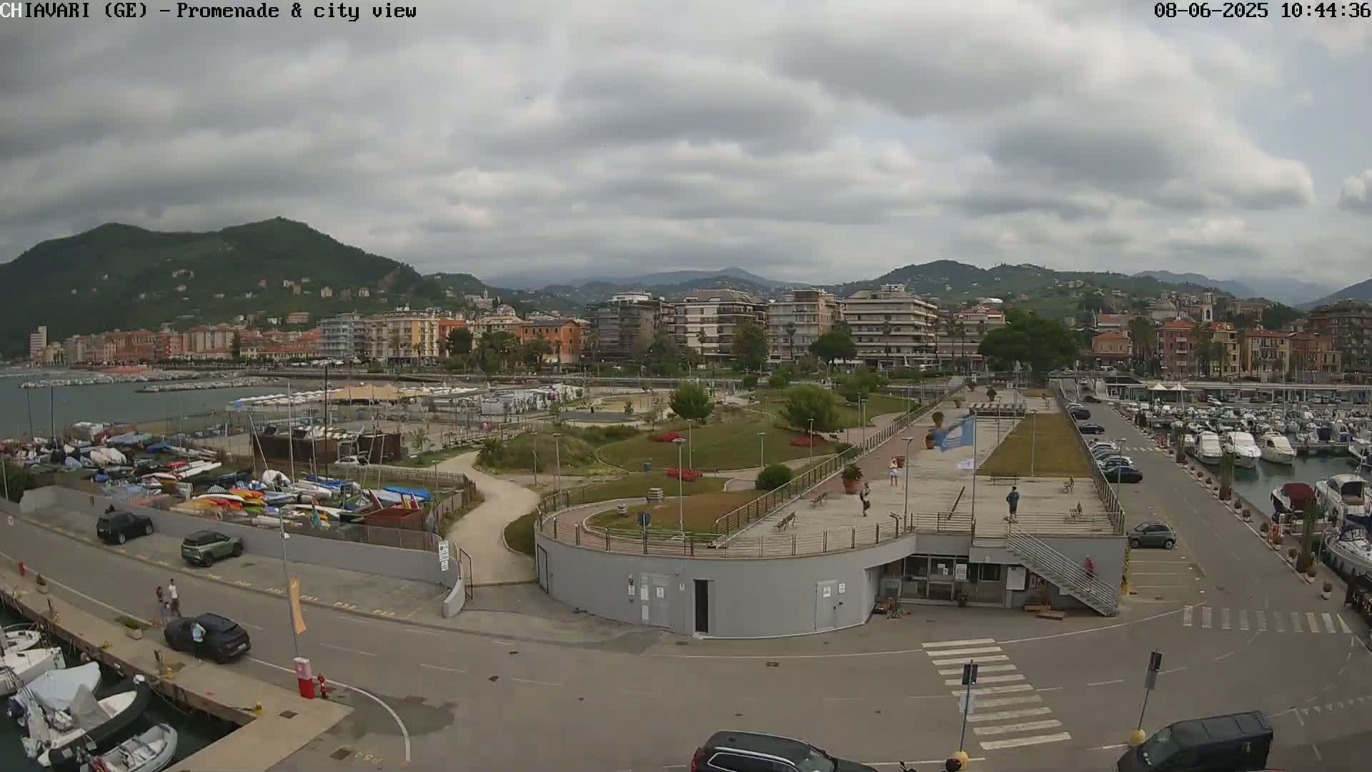 An overcast day reveals a harbor scene with numerous boats, cars parked in a lot, and a landscaped promenade overlooking a town nestled against a mountain range.