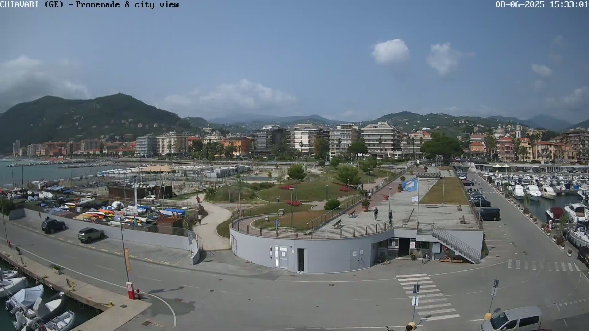 A partly sunny view of a harbor filled with boats, a paved promenade, and a city nestled against mountains in the background.