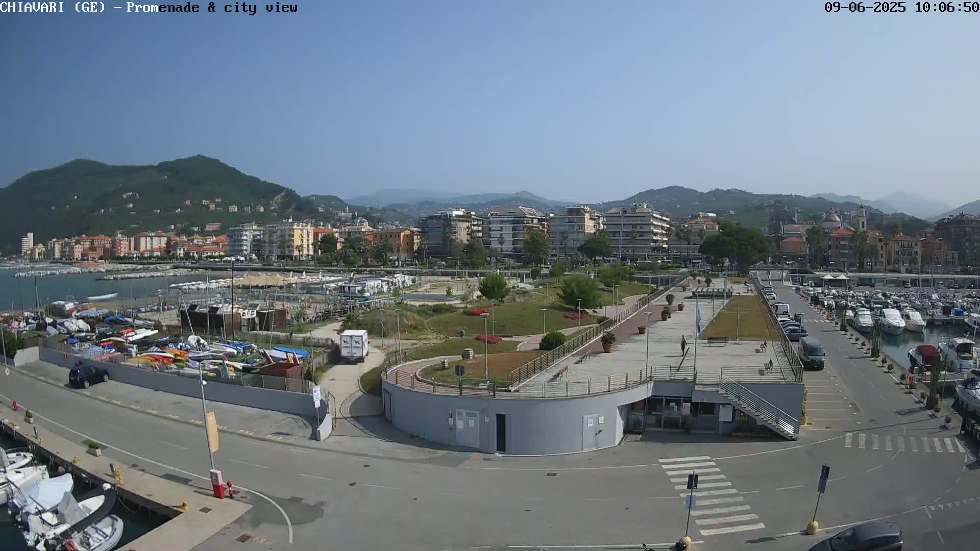 A coastal city scene on a sunny day shows a marina filled with boats, a promenade with landscaping and parking areas, and buildings nestled against a hillside.