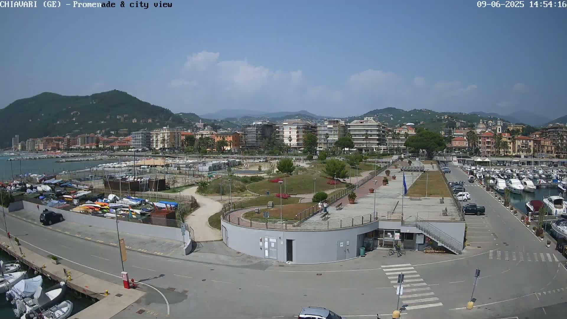 A sunny day reveals a coastal city with a harbor filled with boats, a promenade with landscaped areas, and buildings nestled against a backdrop of hills.