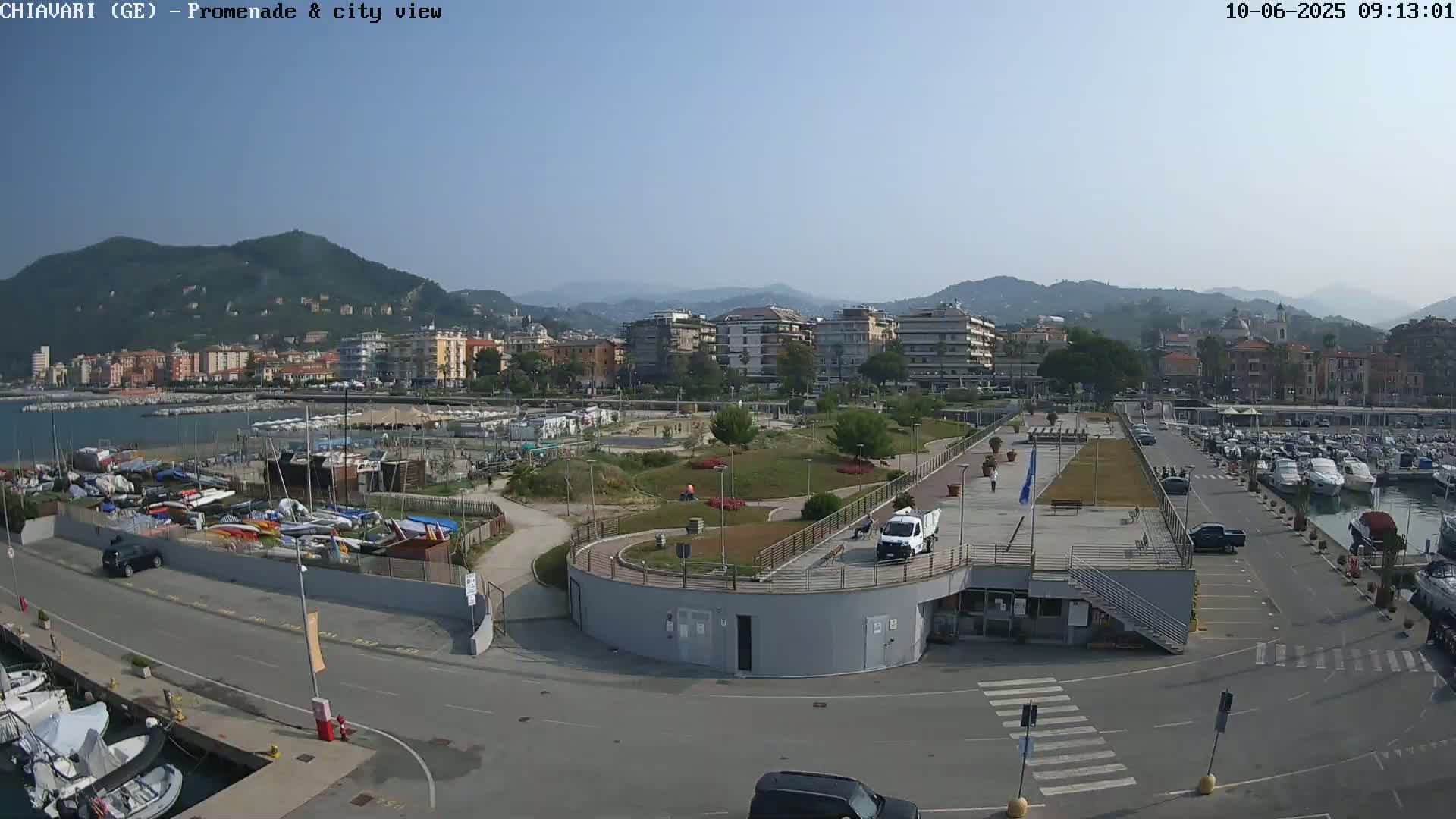 A clear day shows a harbor filled with boats, adjacent to a paved promenade and a city nestled against a mountain range.