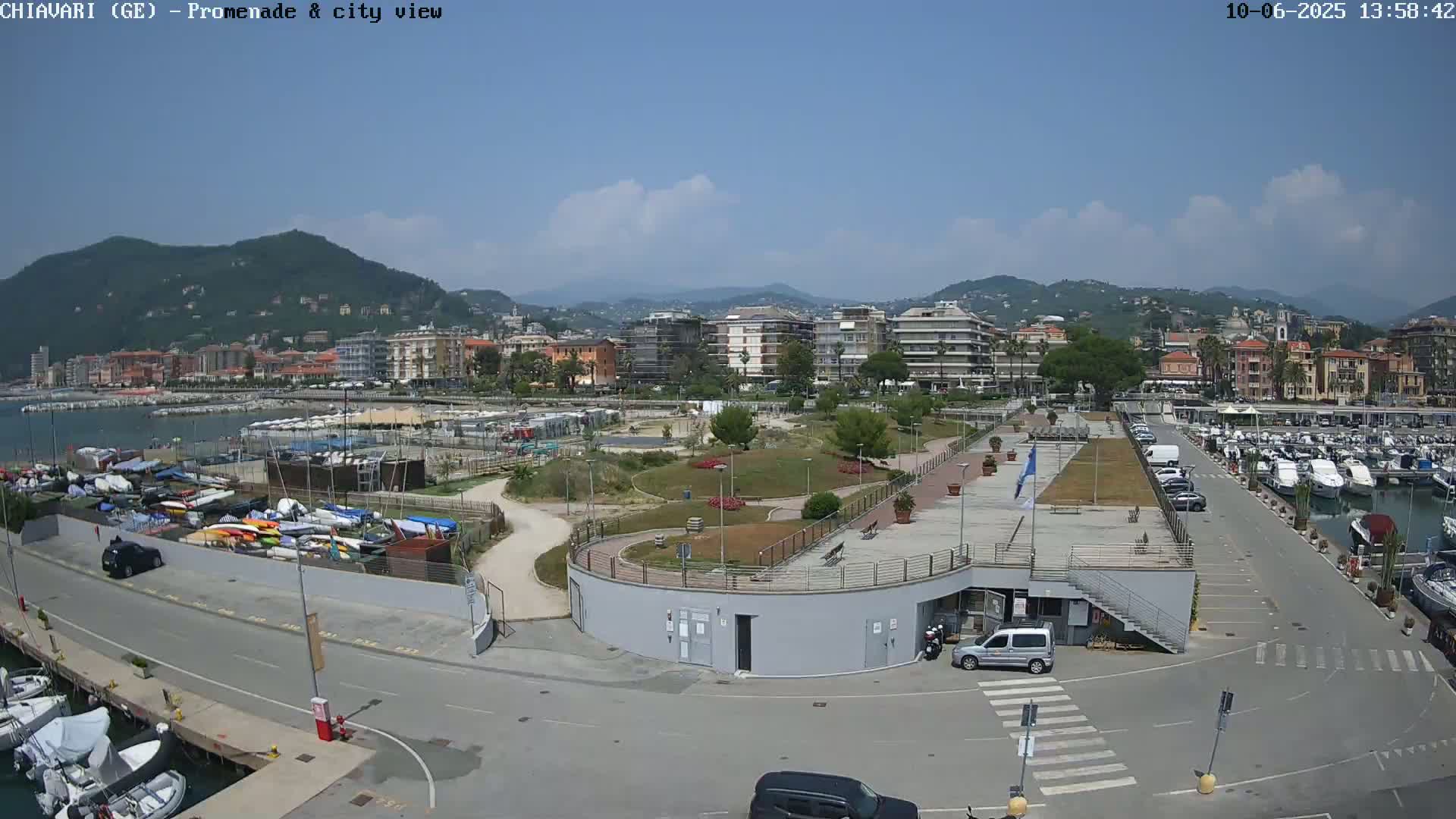 A sunny day overlooking a harbor filled with boats, a landscaped promenade, and a town nestled against mountains in the background.
