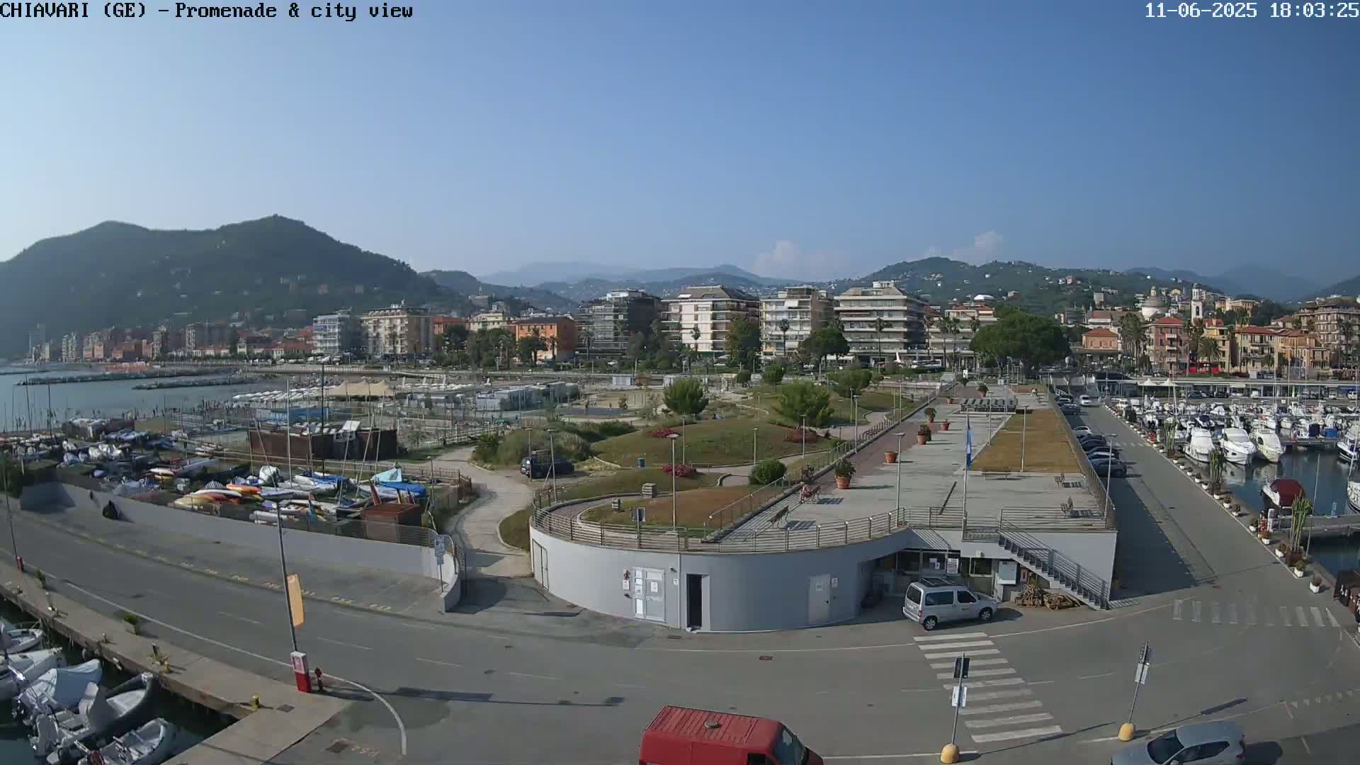 An aerial view shows a harbor filled with boats, a paved promenade, and a coastal town nestled against a mountain range under a clear, sunny sky.