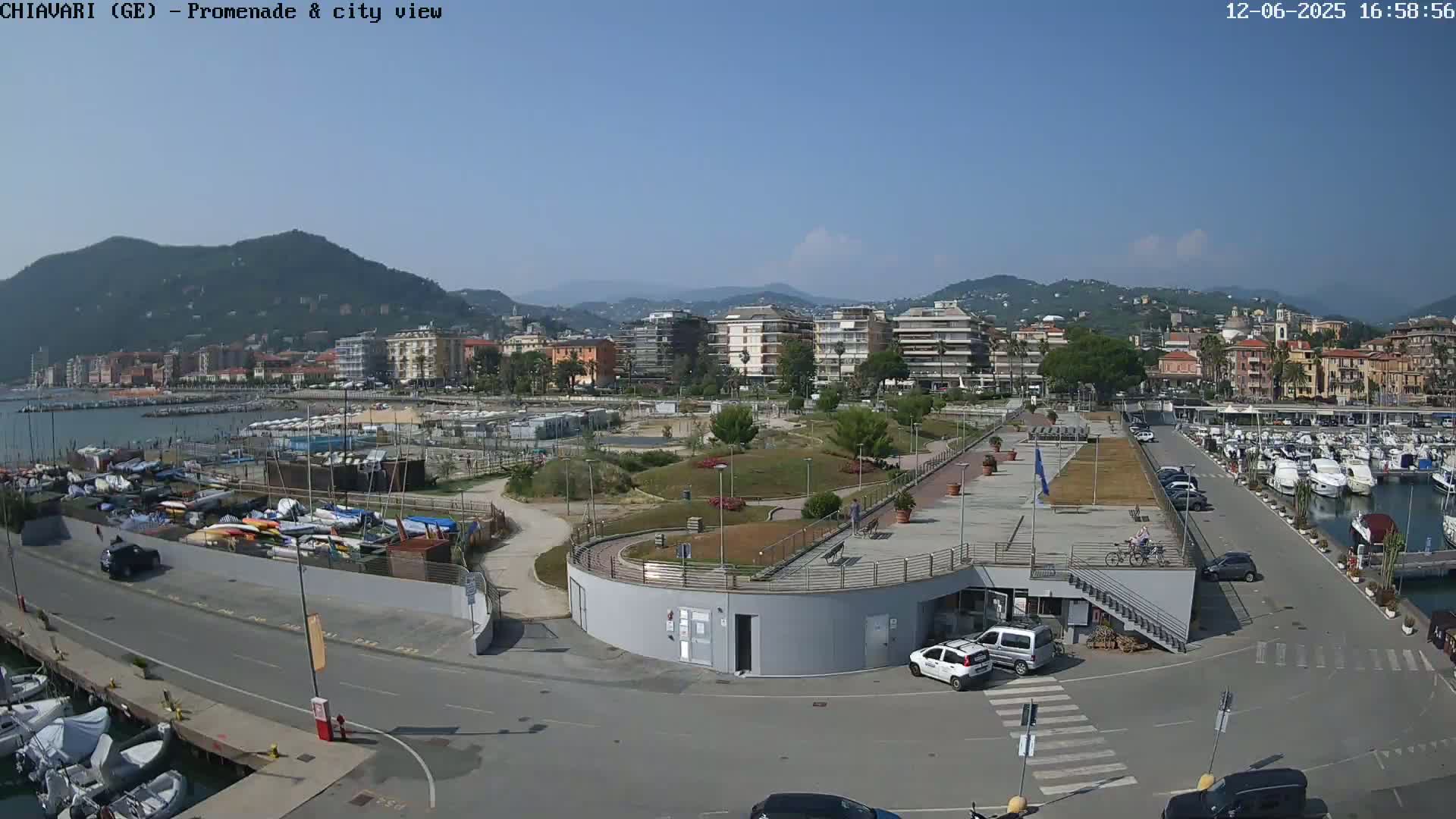A sunny day overlooking a harbor filled with boats, a promenade with a park, and a cityscape set against a backdrop of mountains.