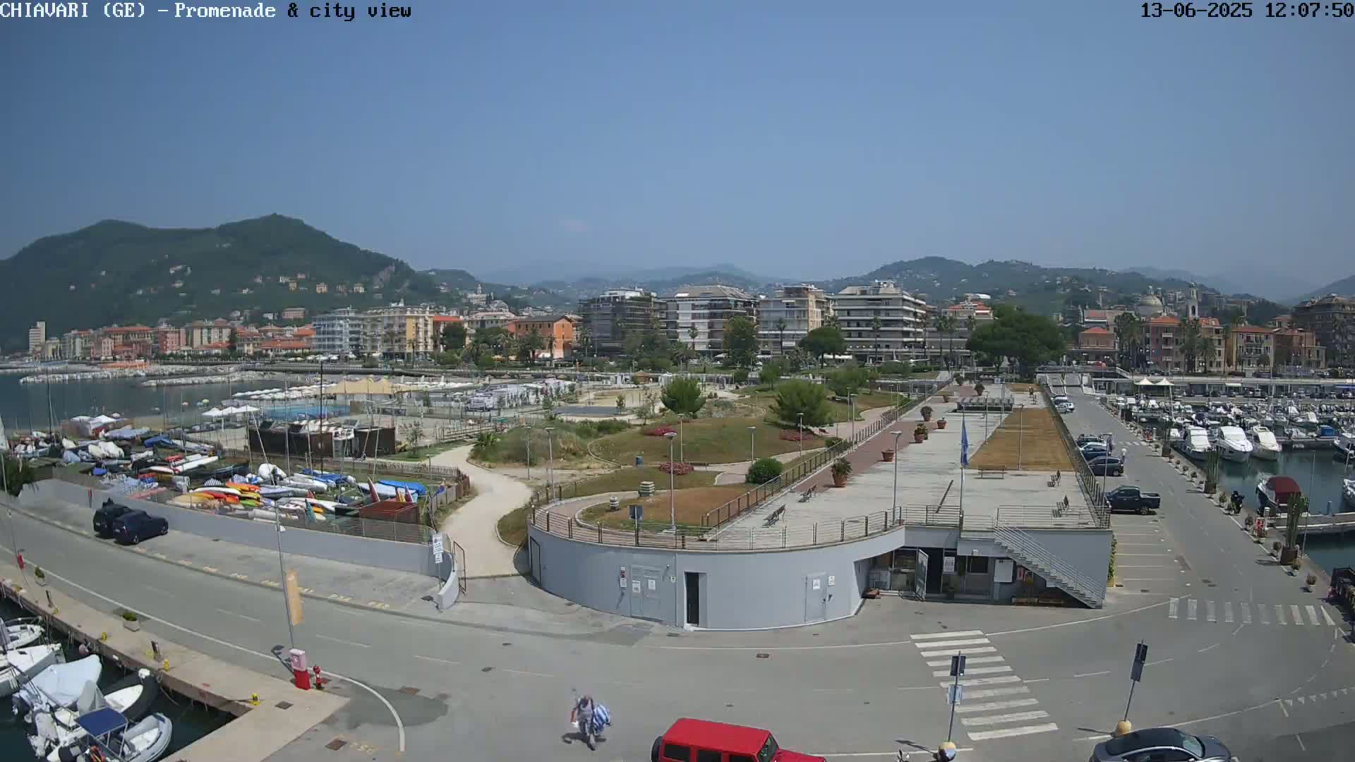 A coastal city scene on a sunny day shows a harbor filled with boats, a promenade with a park, and buildings extending into the hills in the background.