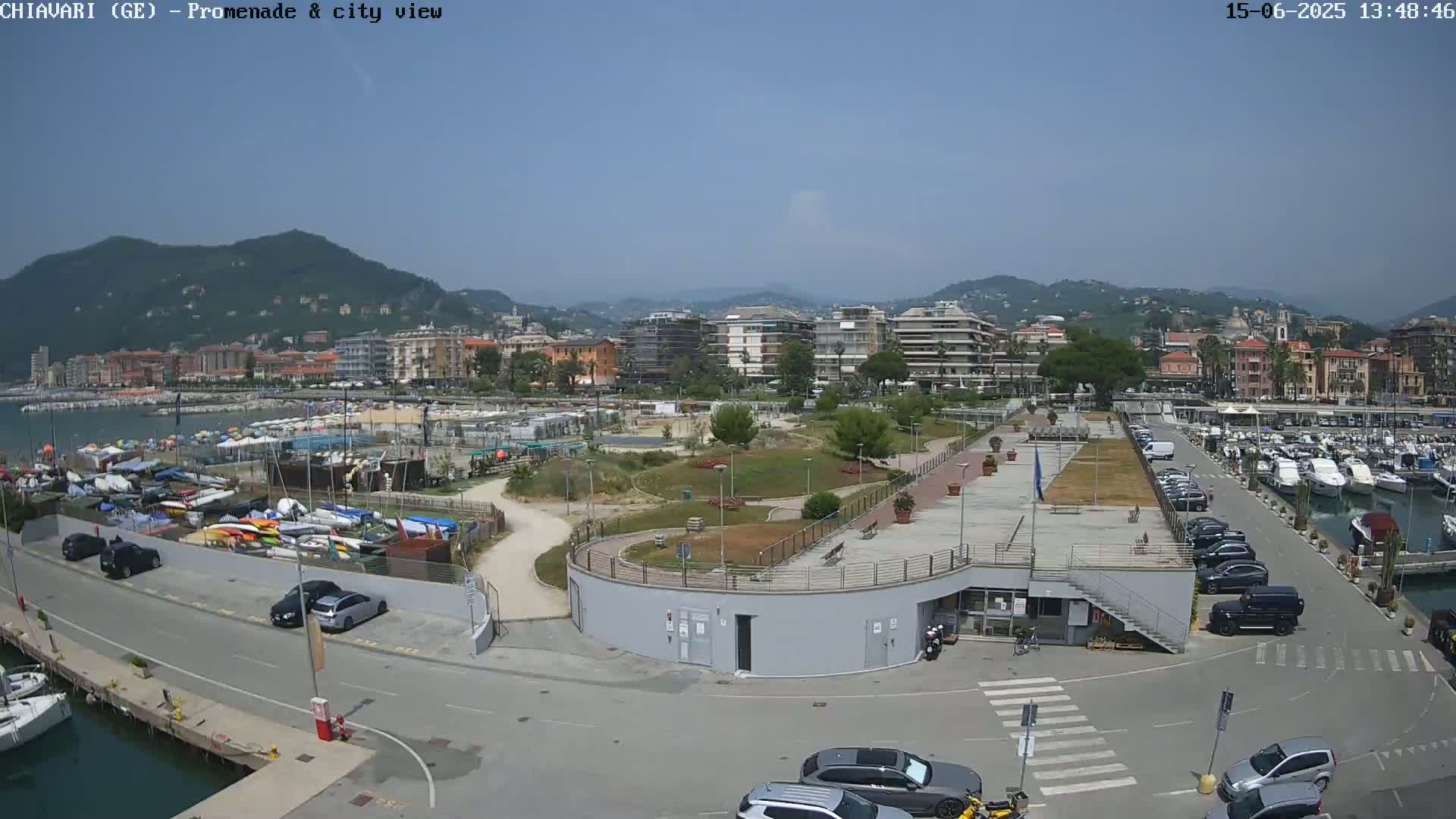 A sunny day overlooking a harbor filled with boats, a promenade, a park, and a city nestled against hills in the background.