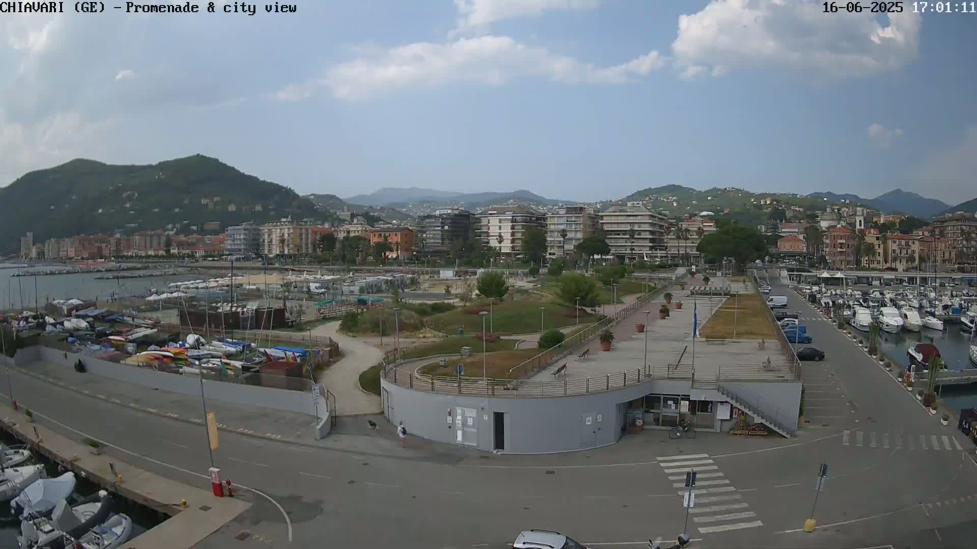 A mostly sunny day overlooking a harbor filled with boats, a waterfront promenade, and a city nestled against hills in the background.